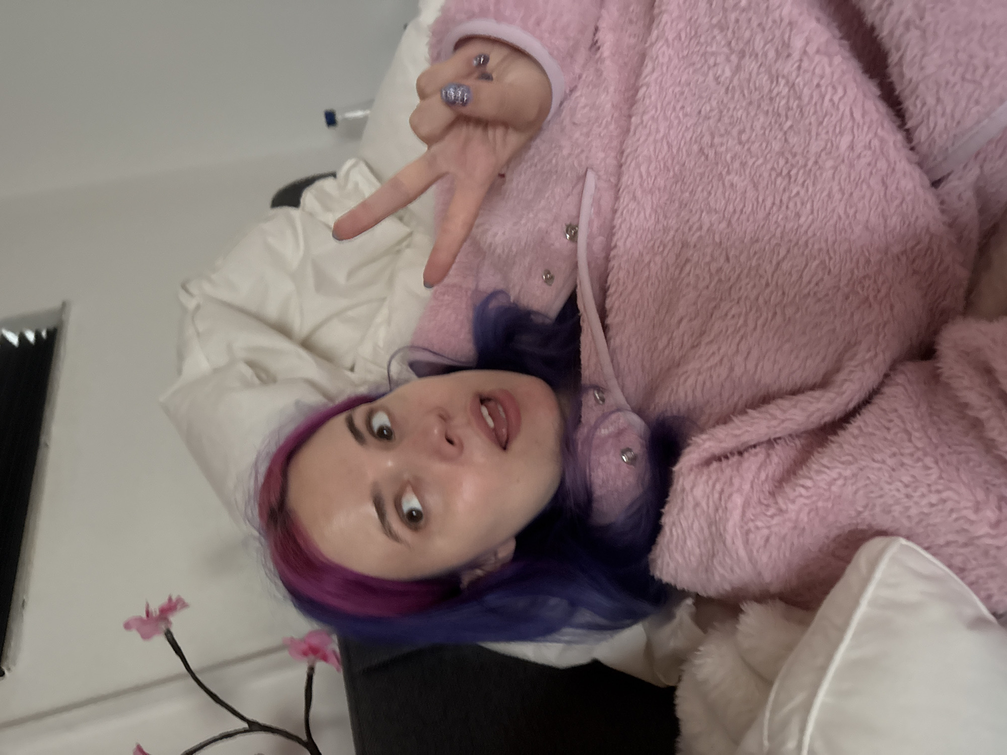 A woman with violet and pink hair is in a pink fuzzy jumpsuit, lying on a bed with white bedding. She is making a peace sign with her right hand. The location is not discernible from the provided image.