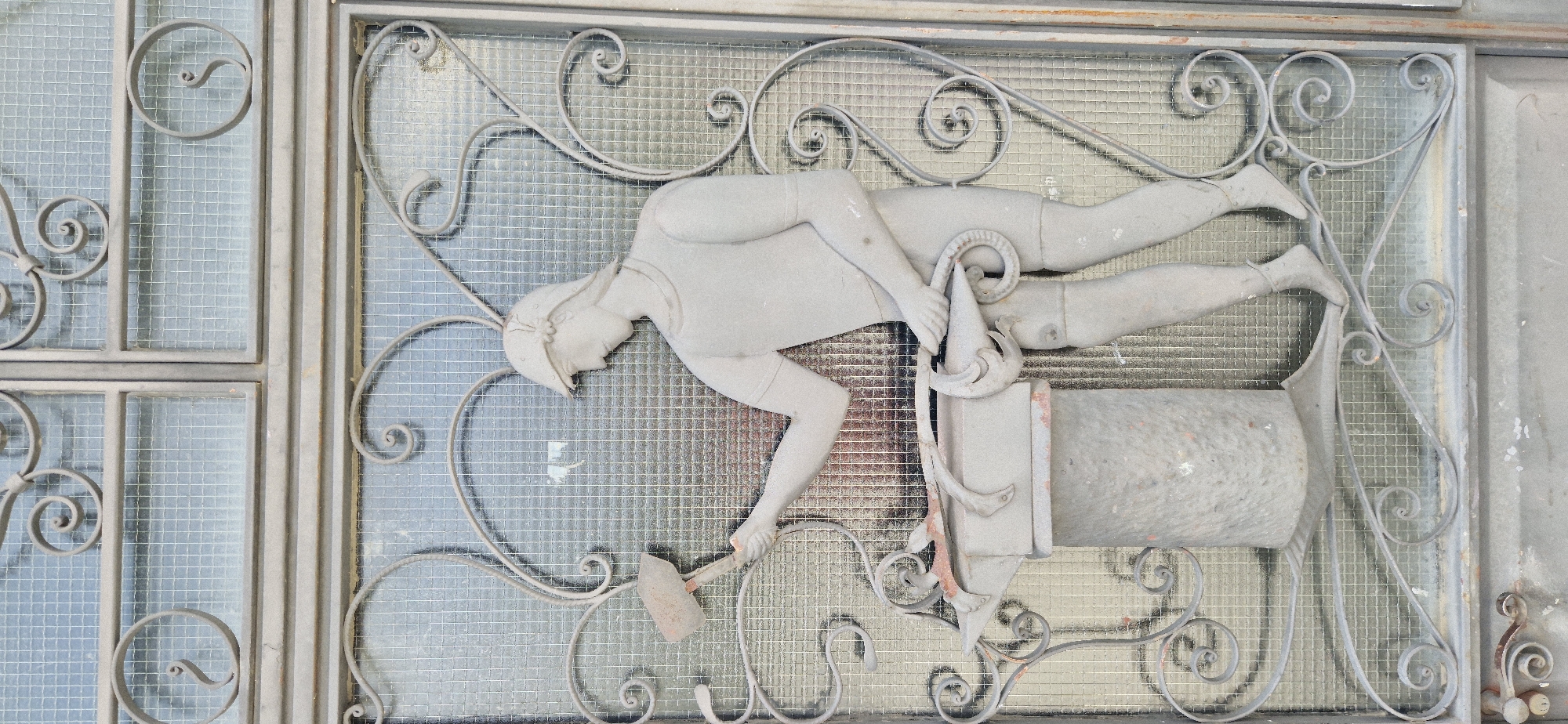 The image displays a wrought iron gate with an embedded relief sculpture depicting a blacksmith. The sculpture is rendered in a light gray material, possibly stone or concrete, and is set against a background of glass panes with a fine grid pattern.
The blacksmith figure, a male, is shown in profile, facing left. He is wearing a cap and a short-sleeved tunic over shorts. His right arm is raised, holding a hammer above an anvil. His left arm is bent, holding a curved metal object, possibly tongs or a piece of hot metal, which rests on the anvil. The anvil itself is depicted as a robust, cylindrical base topped with the characteristic shape of an anvil.
The wrought ironwork surrounding the sculpture features decorative scroll motifs and geometric patterns, typical of decorative gates. The overall scene suggests a classical or historical theme, possibly representing craftsmanship or labor. The lighting is somewhat diffused, highlighting the texture of the relief and the metalwork. The bottom of the image shows the lower part of the gate, including hinges and a latch mechanism. The color palette is dominated by grays and muted metallic tones, with some reddish-brown rust spots visible on the anvil and the lower metal elements of the gate. The environment appears to be outdoors, given the textured glass and the visible elements of the gate structure.