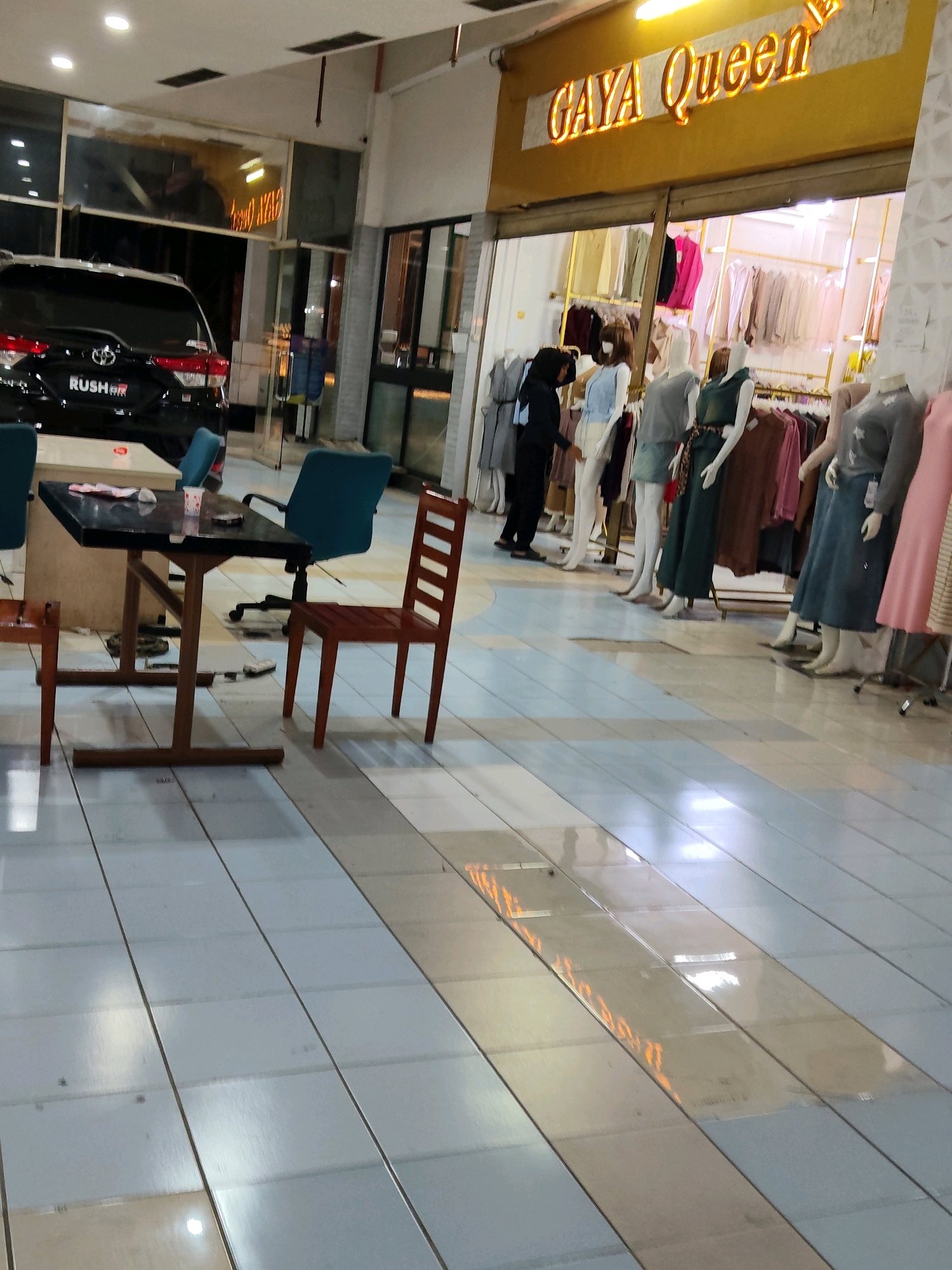 The image depicts an indoor scene, likely a shopping mall or a commercial area in Purwakarta, Indonesia. In the foreground, a dark rectangular table with a glossy surface is situated, accompanied by three chairs: one wooden chair on the right, one blue office chair behind the table, and a partial view of a wooden chair to the left. On the table, there is a small white cup with a red rim, some tissues, and a cell phone.
To the left of the table, a black Toyota Rush GR vehicle is parked, its rear end prominently displayed. Behind the car, a section of a building facade with windows is visible, showing a dark interior and what appears to be illuminated signage with the words "GAYA QUEEN" in golden lettering with a crown emblem.
The environment is characterized by tiled flooring with a repeating pattern of light blue and off-white rectangular tiles. The space is brightly lit by overhead fluorescent lighting, with some reflections visible on the floor. In the background, a clothing store is visible, showcasing mannequins dressed in various garments displayed on racks. Several mannequins, positioned in a line, are dressed in different outfits, including dresses and skirts. A person wearing a dark headscarf and a dark outfit is standing near the mannequins, seemingly interacting with the display. The overall scene suggests a commercial establishment, possibly a boutique or clothing store within a larger shopping complex.