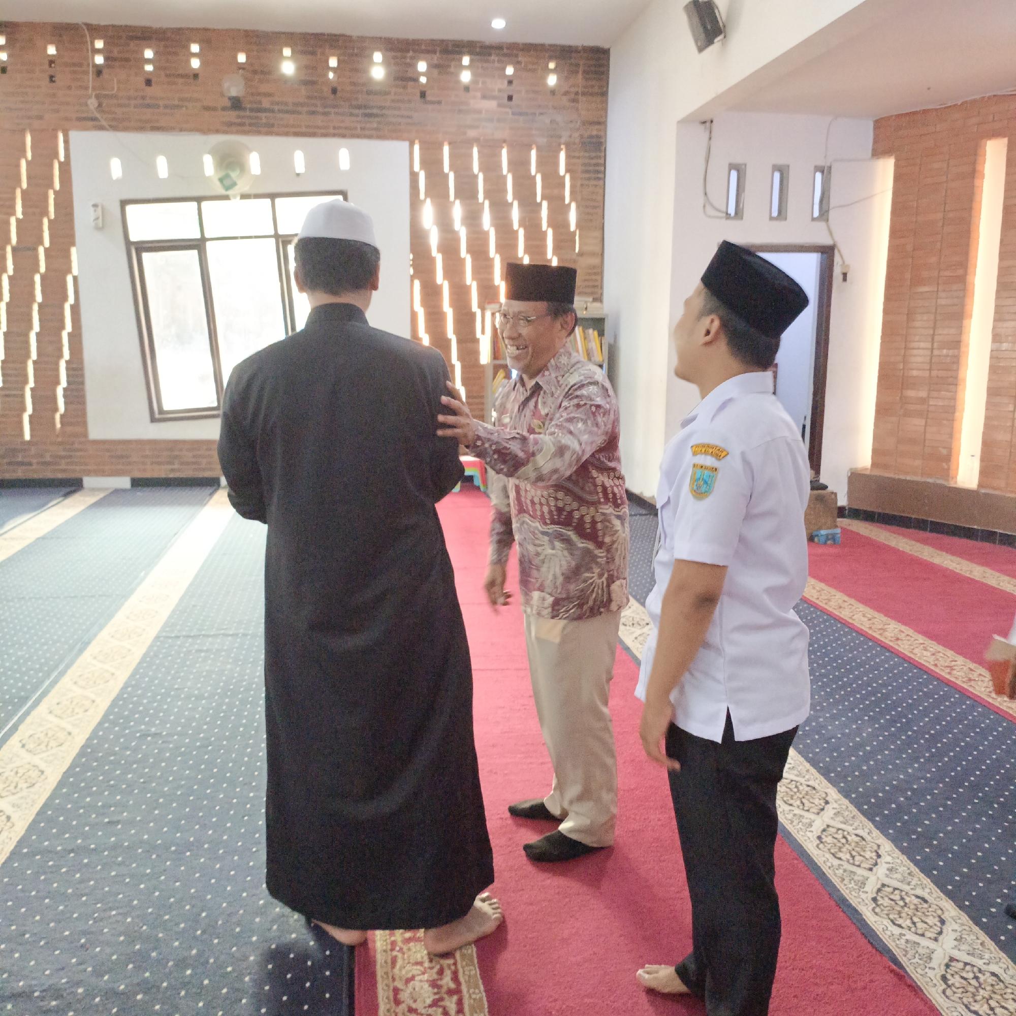 Three men are standing on a red carpeted floor inside a room with blue carpeted sections. The room appears to be a prayer hall or mosque. The man on the left wears a black robe and a white skullcap, facing away from the camera. The man in the center wears a patterned short-sleeved shirt, a cap, and tan pants, and is smiling and extending his right hand towards the man in black. The man on the right wears a white short-sleeved shirt with a logo on the left chest, black pants, and a black skullcap. He is facing the man in the center. The walls are brick and white, with rows of lights running along the upper portion of one wall. There are windows with blinds on the left side of the image, and a bookshelf filled with books is visible in the background. The scene is well-lit, suggesting it is daytime. The presence of prayer mats and the attire of the men indicate a religious or cultural setting. Given the logo on the shirt of the man on the right, which appears to be a government or institutional emblem, and the context of the scene, it is plausible this image was taken in Salatiga, Indonesia, likely at a mosque or Islamic center.