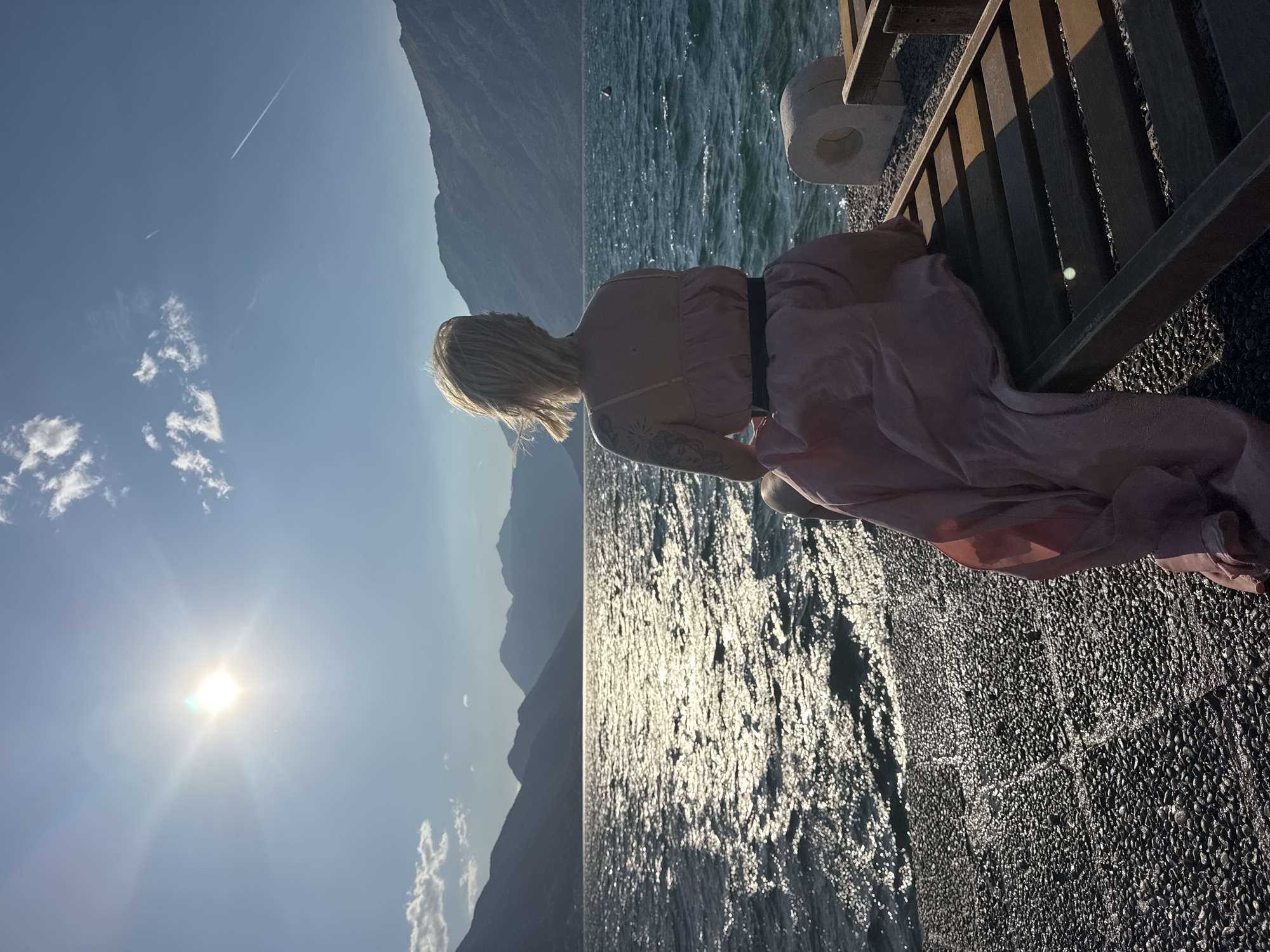 A woman with blonde hair sits on a wooden structure overlooking a body of water with mountains in the background. The woman wears a light pink dress with a dark waistband and has a tattoo on her arm. The sun is bright in the sky with some clouds present. There is also a contrail visible in the sky. The wooden structure the woman is sitting on appears to be a pier or boardwalk. The location is likely Dobrota, Montenegro, given the setting of mountains and water.