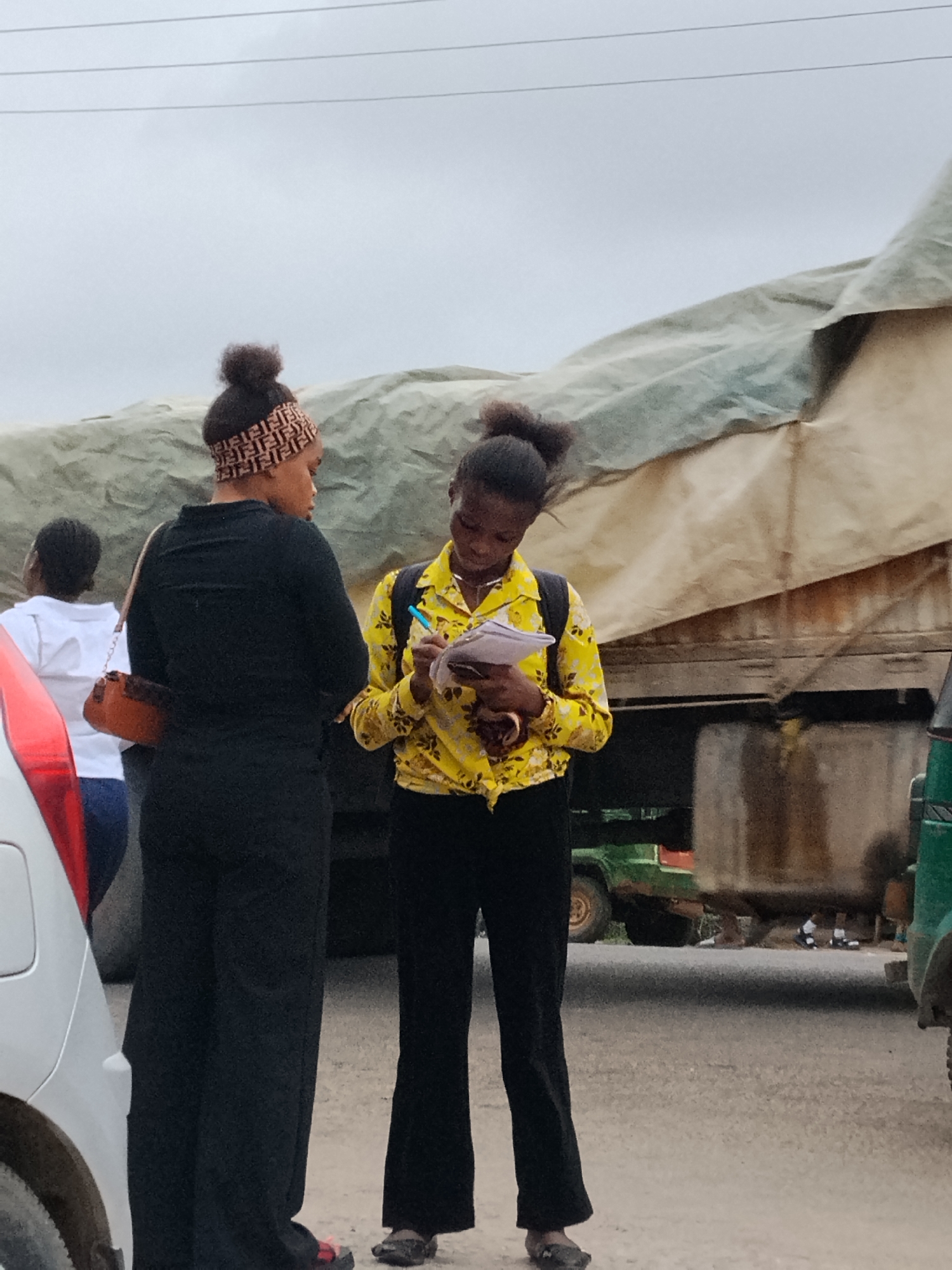 Two young women stand on a street in Aba, Nigeria. One woman wears black pants, a black top, and a Fendi headband, holding a small brown purse. The other wears black pants and a yellow floral shirt, writing on a piece of paper with a pen. Behind them is a large truck covered with a tan tarp, and parts of vehicles are visible on both sides. In the background, other people can be seen near the truck. The sky is overcast. The road surface appears to be paved.