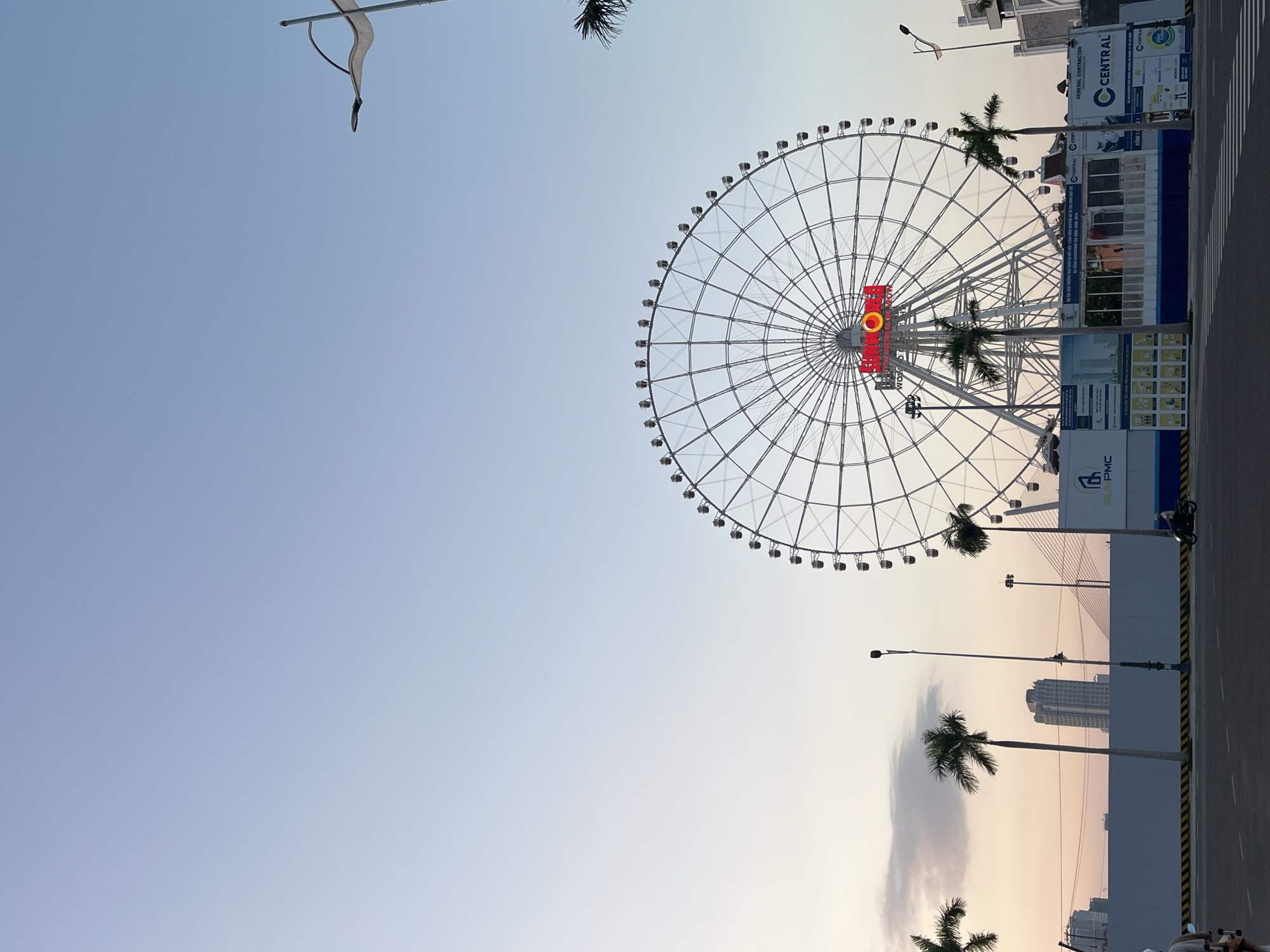 The image, oriented vertically, captures a large Ferris wheel dominating the right side of the frame against a clear, gradient sky. The sky transitions from a light blue at the top to a warm, pale orange and yellow closer to the horizon, suggesting either dusk or dawn.
The Ferris wheel is white or silver with numerous passenger gondolas evenly spaced around its circumference. A prominent, illuminated red and yellow logo at the center of the wheel clearly displays the text "SUN WHEEL".
Several tall palm trees are visible, some partially cropped, adding a tropical element to the scene. To the right of the Ferris wheel, there appears to be a construction or temporary site, marked by white barriers with yellow and black striped safety tape. Behind these barriers are various structures and large signs. One sign prominently features the logo "C CENTRAL" and the text "RENTAL CONTRACTOR", with other smaller text possibly in Vietnamese. Another sign bears the initials "PMC". A multi-story building is visible in the background beyond these structures. A street or road runs along the right side, and a motorcycle is partially visible behind the barrier.
The scene depicts a calm, static view of an entertainment area, likely a park or complex, in an urban or semi-urban environment. The time of day, indicated by the sky's colors and the illuminated Ferris wheel logo, is most likely evening. The overall weather is clear with minimal cloud cover. The specific location is implied to be Đà Nẵng, Vietnam. No specific human activity or event is taking place.