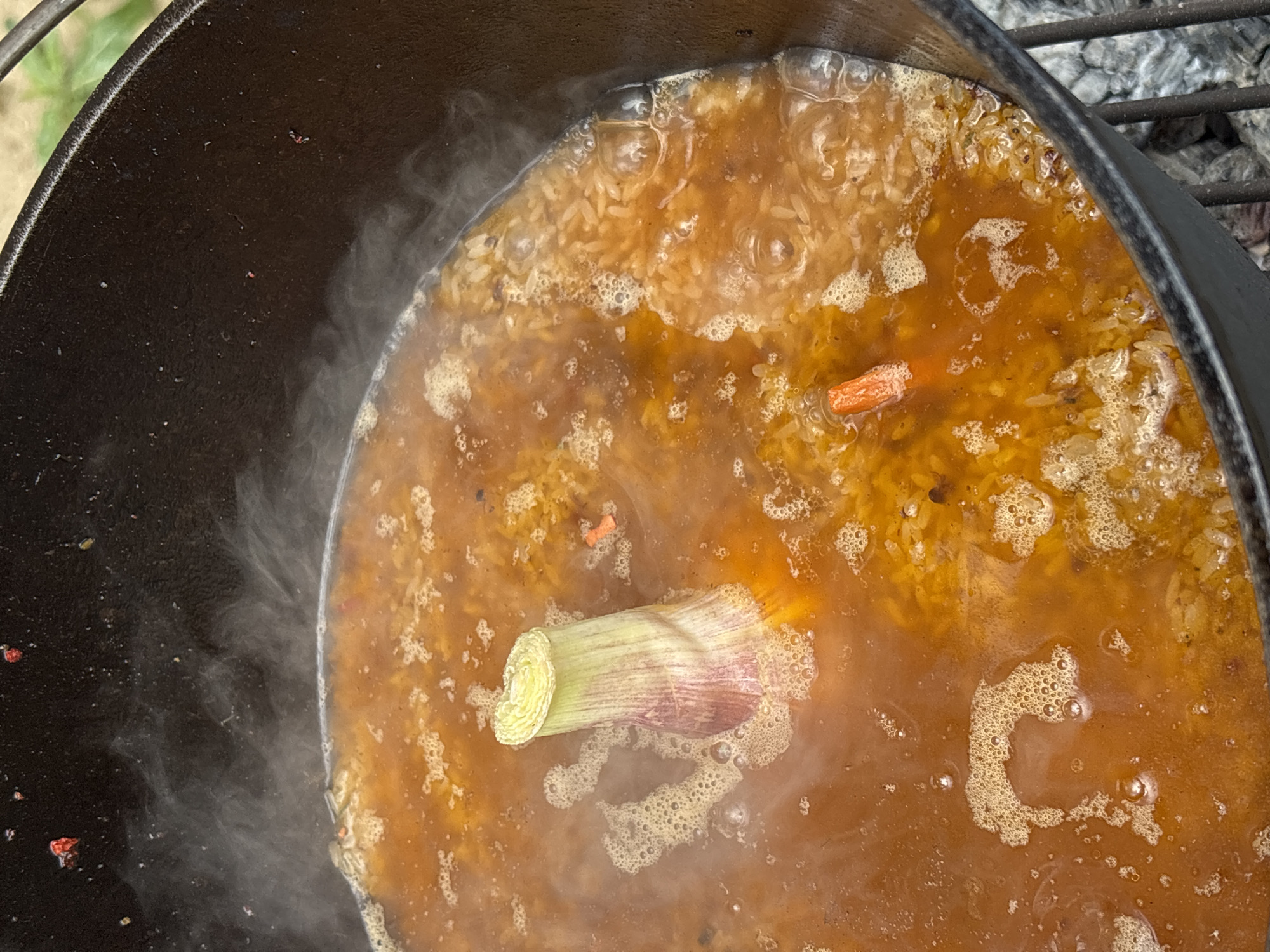 A top-down close-up view of a dark brown or reddish-brown liquid simmering in a black cast iron pot. A section of leek is prominently placed in the middle of the pot. Smaller pieces of carrots and rice are visible in the simmering liquid. Steam rises from the side of the pot. The scene is situated on an outdoor cooking setup with a metal grill visible on the side of the pot, above embers of a fire that heat the pot. It is difficult to pinpoint the location as Prikamskii, Russia, without additional contextual clues.