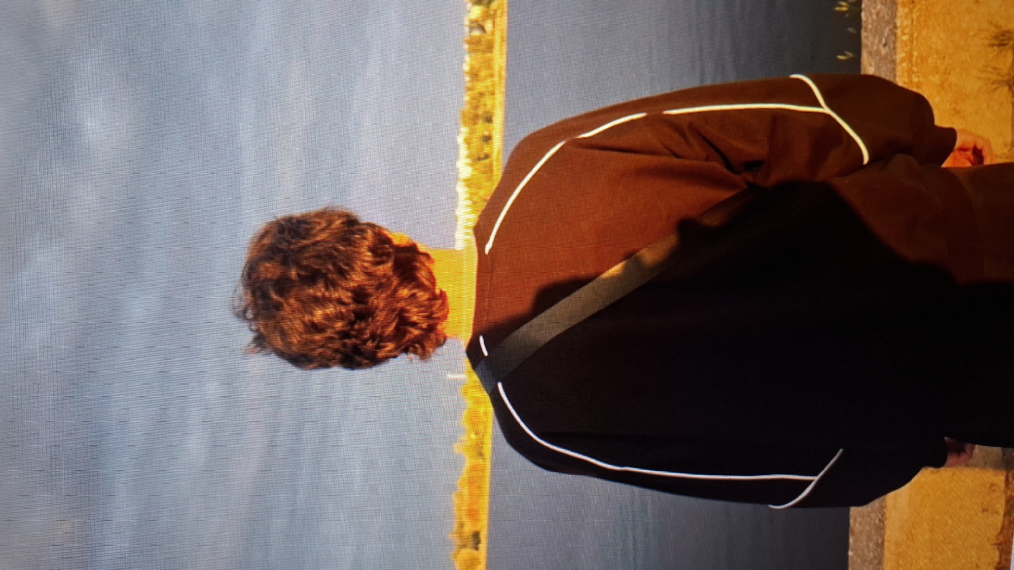 The image shows the back of a person with curly, reddish-brown hair. They are wearing a dark jacket with white piping and a dark band across the back. The person is bent over, obscuring their face. The background is a blurry expanse of blue, possibly water or sky, with a bright yellow-orange vertical stripe to the left of the person. The lighting suggests it might be daytime, perhaps late afternoon or early morning, given the warm tones in the yellow-orange stripe. The overall mood is somewhat ambiguous due to the obscured subject. There is no visible text in the image.