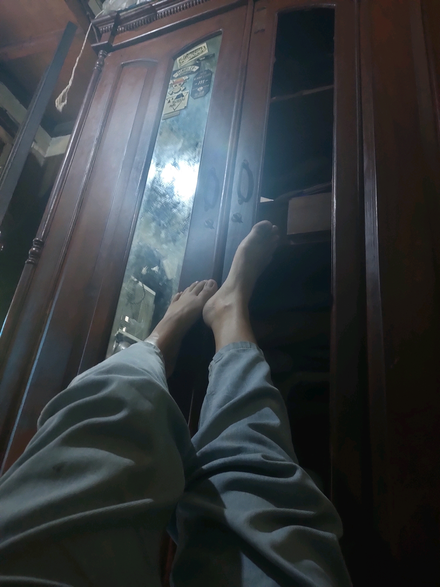 The image depicts a low-angle view of a person's legs and feet resting against a dark wooden wardrobe in what appears to be a room. The wardrobe has a mirrored door that reflects some light and the environment, including what looks like a window or light source. On the mirrored door, several stickers are visible, including one with text that appears to read "Glimmers." The person is wearing light-colored, loose-fitting pants. One door of the wardrobe is slightly ajar, revealing an empty shelf inside. The lighting is dim, suggesting it may be nighttime or the room is not well-lit. The overall impression is one of relaxation or repose. The location is specified as South Jakarta, Indonesia.