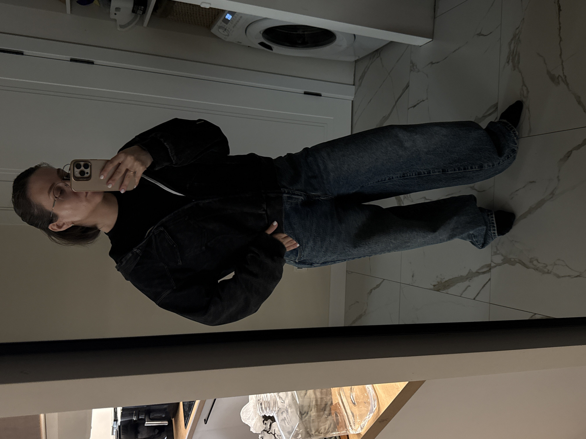 A full-body mirror selfie captures a person standing upright in an indoor setting. The person, likely female, has dark hair pulled back and wears glasses. They are dressed in a dark denim jacket over a dark top and loose-fitting, light-blue jeans. Their right hand holds a light-colored smartphone, displaying white nail polish on their fingers. A ring is visible on the ring finger of their left hand, which is placed on their hip.
The background, reflected in the mirror, reveals a domestic interior. Above the person's shoulder, a white washing machine is visible, featuring a digital display showing the number "28". Below the washing machine, there is a white door or cabinet. The floor is covered with white marble tiles accented with grey veining. Below the dark frame of the mirror, a reflection shows a wooden surface and several transparent objects, possibly glass bottles or vases.
The person is engaged in the activity of taking a self-portrait. The indoor lighting appears somewhat dim, suggesting it's not bright daylight. The presence of the washing machine and tiled floor indicates a utility room or bathroom, fitting for a residence in Pervomaysky, Russia. No other discernible text is visible besides the number "28" on the appliance.
