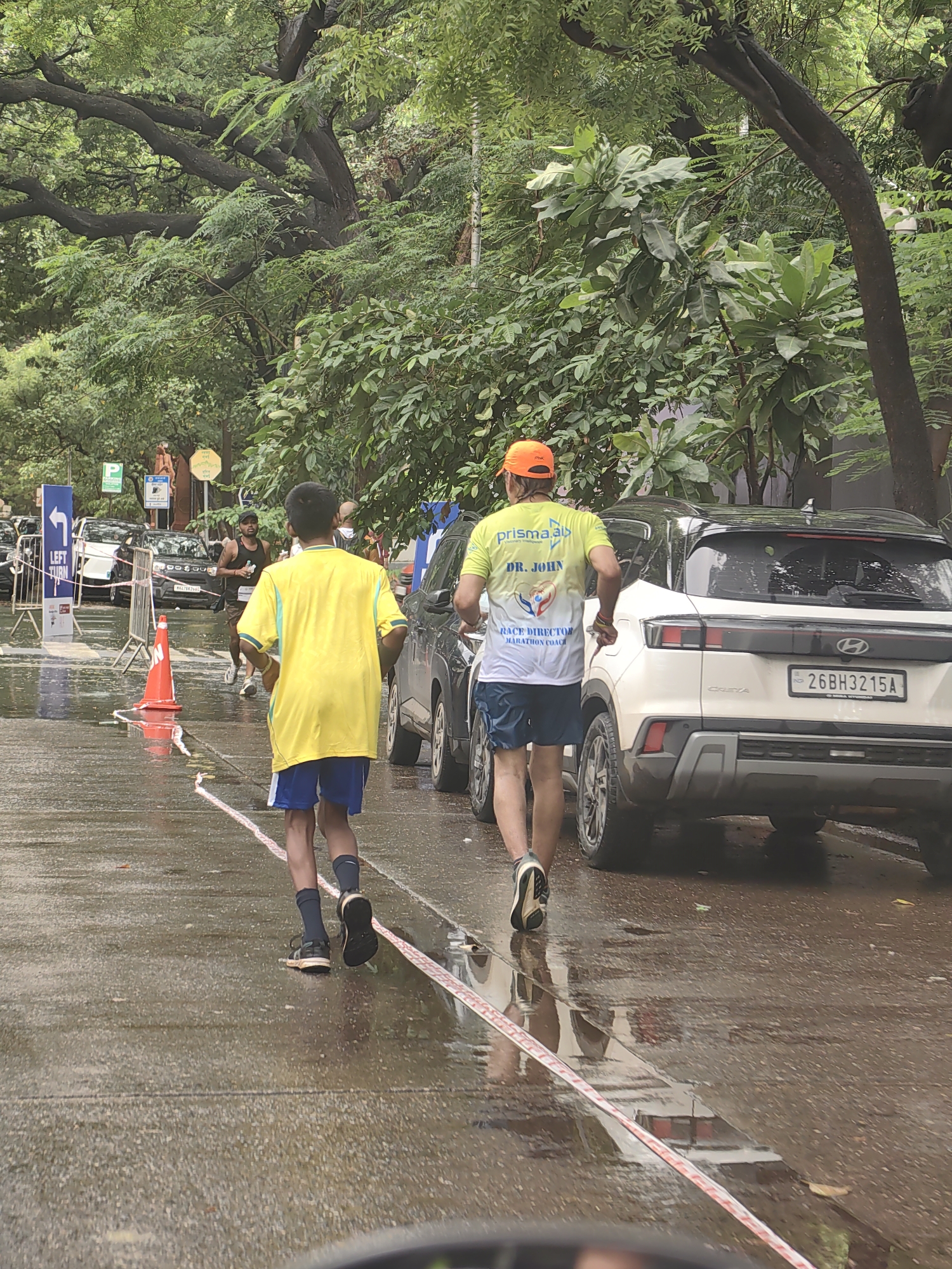 The image shows a marathon scene on a wet road in Mumbai, India. Two male runners are visible in the foreground, one in a yellow shirt and blue shorts, and another in a light green shirt with "DR. JOHN" and marathon-related text on the back, wearing an orange cap. Several cars are parked or driving on the road, including a white Hyundai Creta with a license plate ending in "3215A". Traffic cones and barriers are set up along the racecourse. Lush green trees line the sides of the road, indicating an urban park or tree-lined street. The road surface is wet, suggesting recent rain or a washed course. Signs indicating "LEFT TURN" are also visible, suggesting a street intersection. The overall atmosphere appears to be that of an organized outdoor event amidst a slightly overcast or post-rain environment.