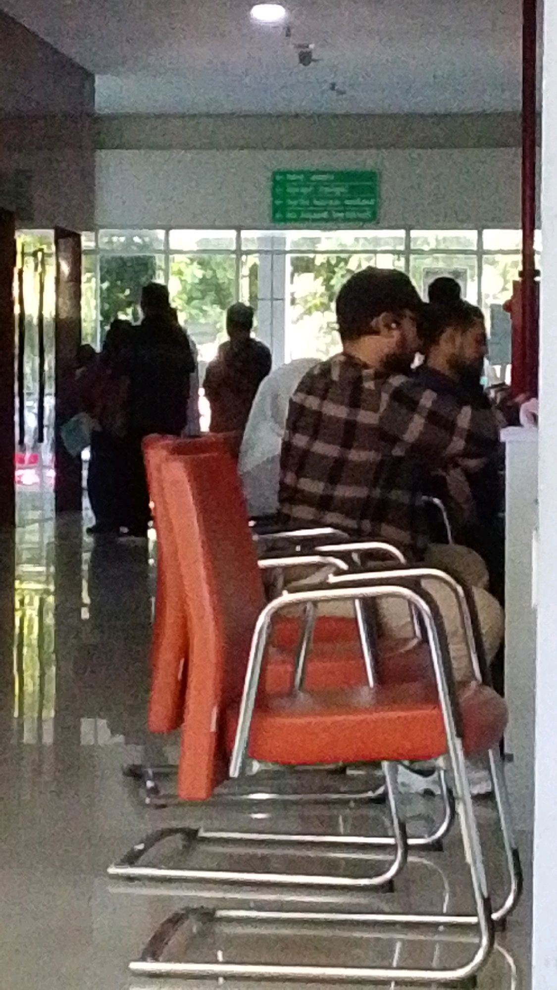 In Pabelan, Indonesia, an indoor scene depicts a waiting area, likely within a public building. The foreground is dominated by two chairs, one a bright orange plastic seat with a metal frame, and another of similar construction behind it. The seated subjects are men, one in a dark and light plaid shirt, and another partially visible. A few other individuals are present in the midground, blurred and indistinct, some standing. The background reveals large windows looking out onto greenery, suggesting a connection to the outdoors. A green sign with text in an unknown language is affixed to the wall above the windows. The overall impression is of a functional, everyday space for waiting.