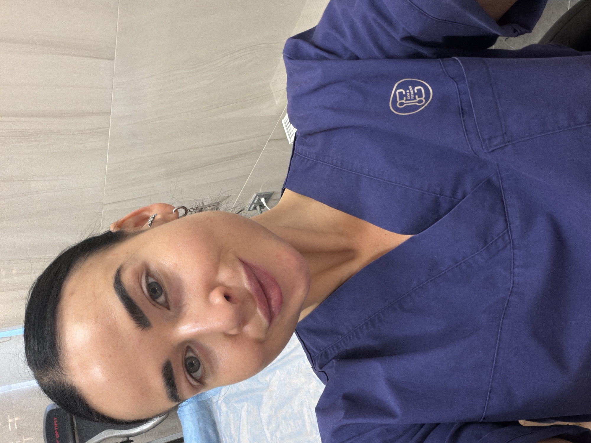 A woman in a dark blue medical or work uniform is positioned within a room. The background includes light-colored tiled walls and what appears to be medical or examination equipment covered with a light blue sheet. The woman's uniform has an emblem that may signify "Gym & Fitness." The presence of medical equipment suggests the setting may be a clinic or similar medical facility. Without any additional information, determining if this is in Saint Petersburg, Russia, is impossible.
