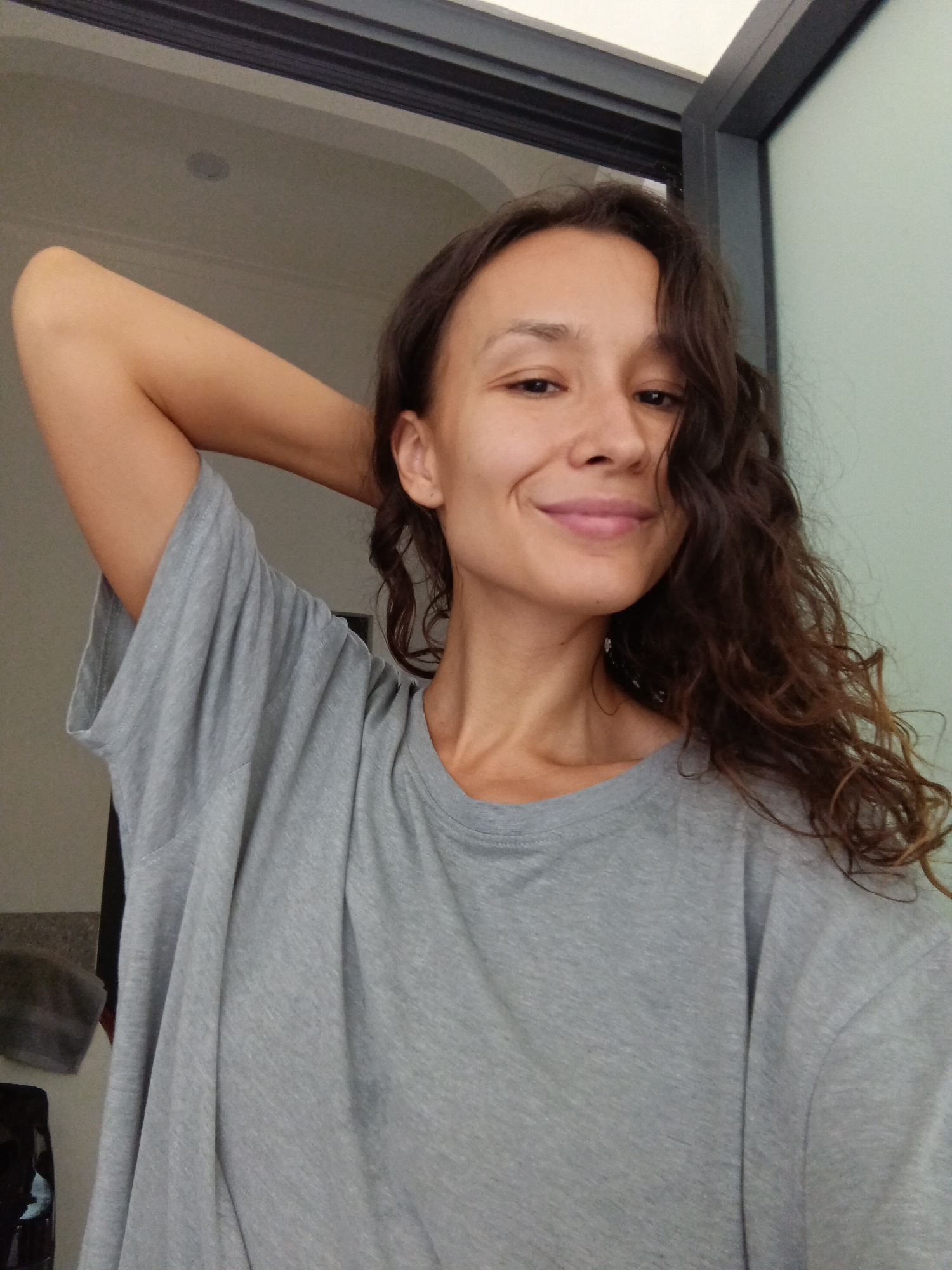A young woman with curly brown hair smiles at the camera with her right arm behind her head. She is wearing a plain gray t-shirt. The background is blurred and appears to be indoors, with a doorway visible to the right. There is no indication of the city of Mũi Né, Vietnam in the image.