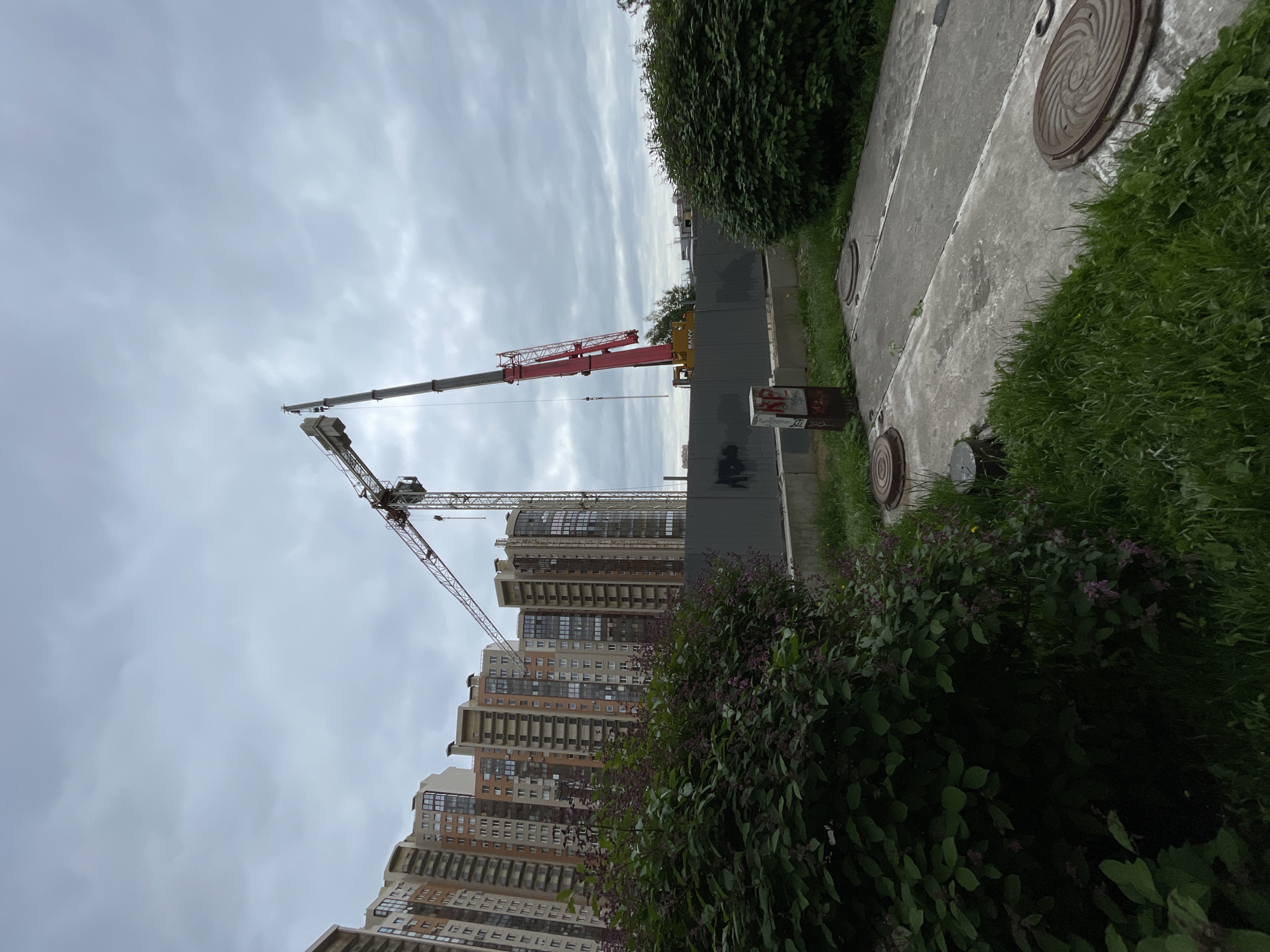 The image depicts a construction site in Saint Petersburg, Russia. Two cranes are visible, one red and one grey, both extending over partially constructed high-rise apartment buildings. A concrete sidewalk runs along the edge of the construction site, bordered by dense green bushes. Several manhole covers are visible on the sidewalk. The sky is overcast with grey clouds. A section of a grey concrete wall is present, possibly acting as a barrier for the construction site. The buildings appear to be residential, with multiple stories and windows.