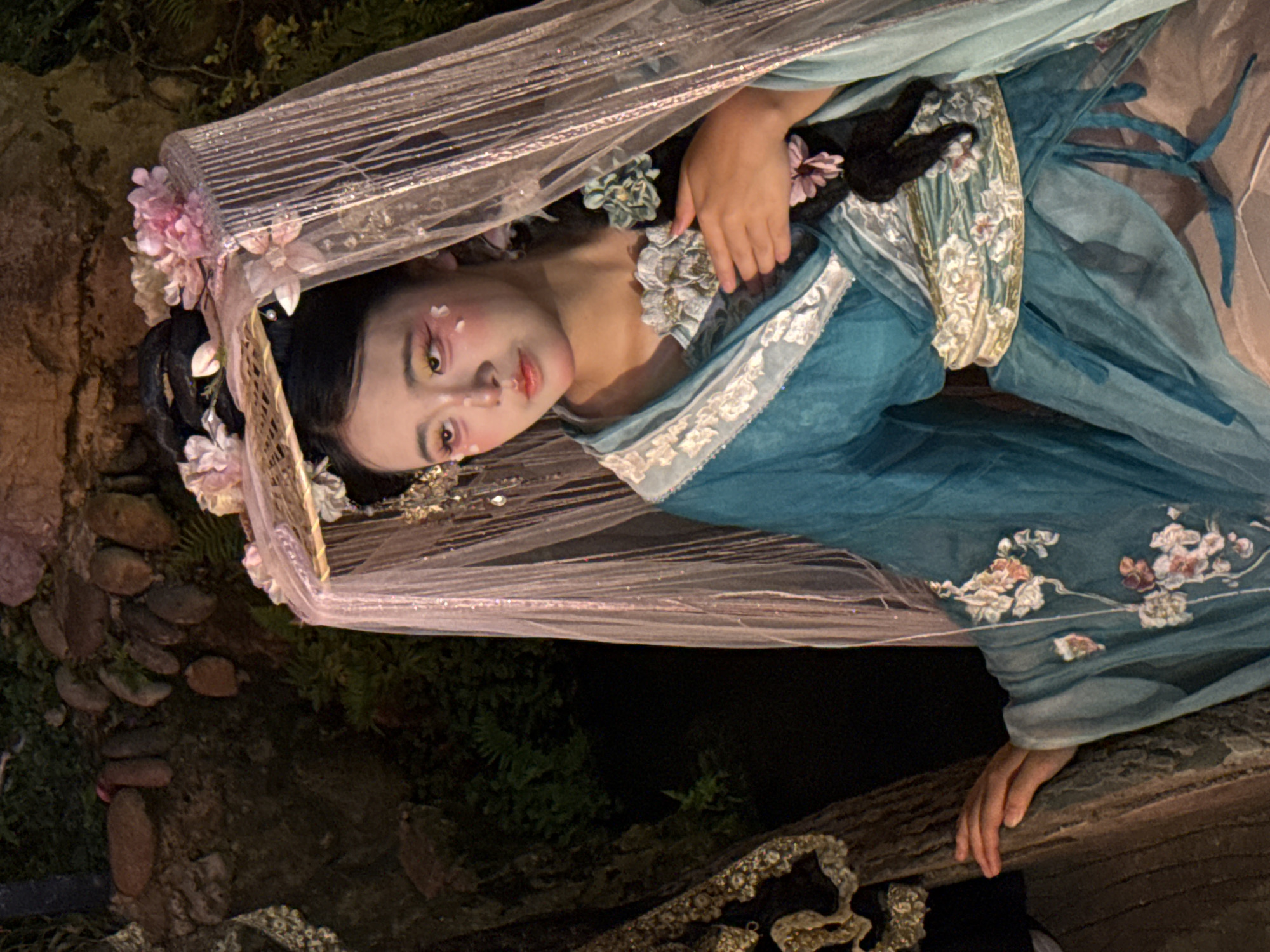 A young woman, dressed in traditional Chinese attire, reclines under a sheer, sparkling veil. Her elaborate makeup, featuring pink accents around her eyes and on her cheeks, evokes a delicate, ethereal beauty. The blue silk robe she wears is adorned with intricate floral embroidery in shades of pink, peach, and white, highlighting the artistry of the garment. Her dark hair is styled with floral accessories, further enhancing the traditional aesthetic.
The woman is positioned within a natural setting, with hints of greenery and textured stones visible in the background, suggesting an outdoor scene. She rests her hand on a wooden surface, which appears weathered and adds a rustic element to the composition. The overall mood is serene and contemplative, with the soft lighting and diffused veil contributing to a dreamlike atmosphere. The scene evokes a sense of artistry and cultural representation, possibly within the context of a performance or a themed photoshoot. There is no visible text in the image.