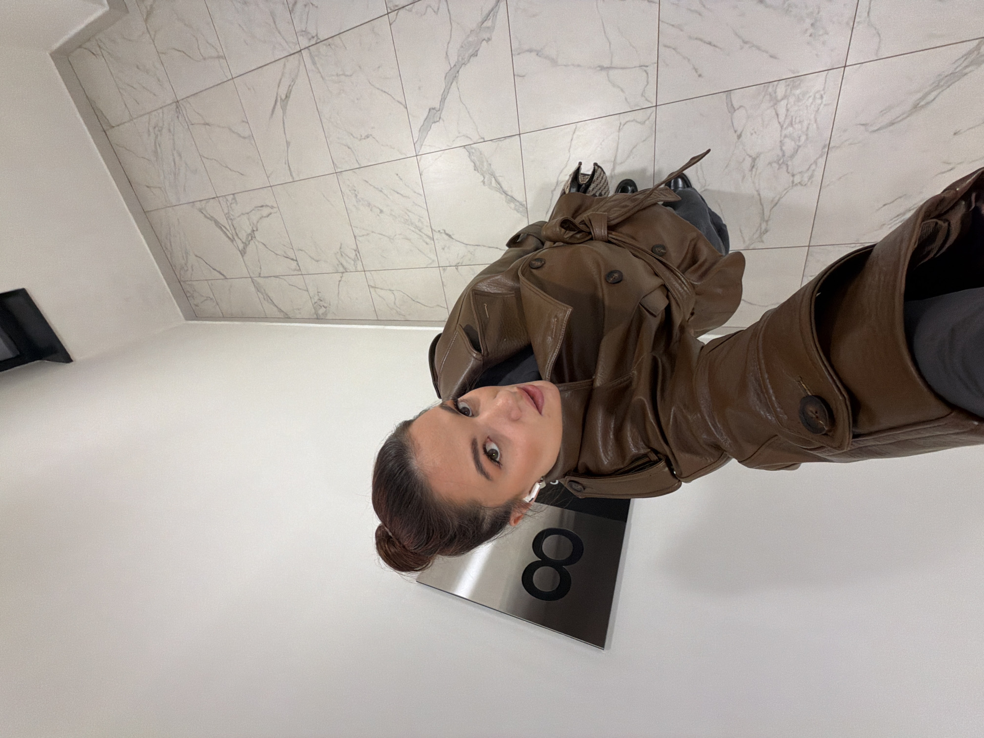 A woman is depicted from a high-angle perspective, consistent with a selfie, positioned within an interior hallway or lobby. She wears a brown, double-breasted trench coat with a belted waist and large dark buttons. Her dark hair is pulled back into a bun, and she has white wireless earbuds in her ears. She looks directly at the camera. The immediate environment includes plain white walls and a white marble-patterned tiled floor with grey veining. On the white wall, to the left of the woman's head, a rectangular metallic plaque is affixed, displaying a prominent black number "8" with a black infinity symbol (∞) directly above it. A dark, rectangular fixture is partially visible at the upper right ceiling line. This location is identified as the Infinity Belgrade Business Center, situated at Bulevar Milutina Milankovića 11A, Novi Beograd, Belgrade, Serbia.