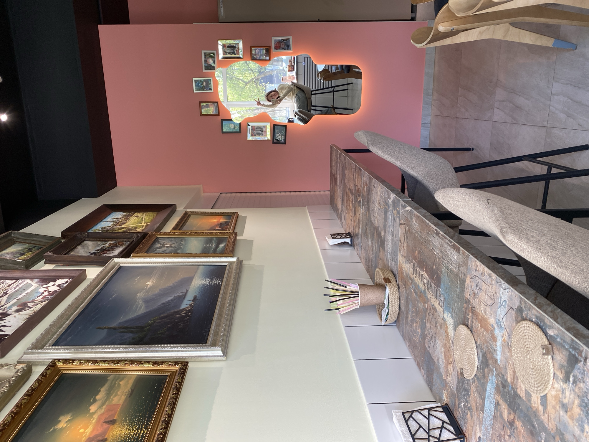The image depicts the interior of a restaurant in the city of Chaikovsky, Russia. The restaurant features a salmon-colored wall with a decorative mirror surrounded by small framed pictures. The reflection in the mirror shows a person inside the restaurant giving a peace sign. The wall is decorated with various landscape paintings, including seascapes and nature scenes. A long counter with stools is positioned along the side, with straw placemats and a straw container filled with straws.