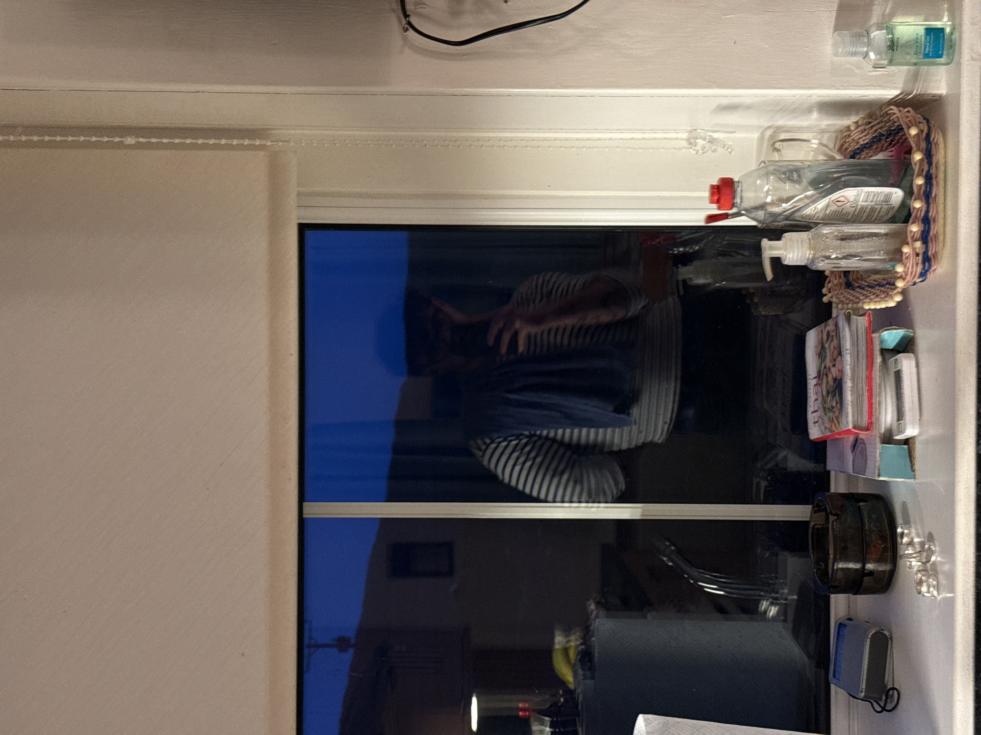 The image appears to be taken indoors, looking through a window. The reflection in the window shows a person wearing a striped shirt, potentially taking a photo. Outside the window, blurred shapes suggest buildings or other structures. The window ledge has several objects on it, including bottles of hand sanitizer, books, a container, earrings, and a blue electronic device. A cord is plugged in at the top of the window frame. The location is currently unknown.