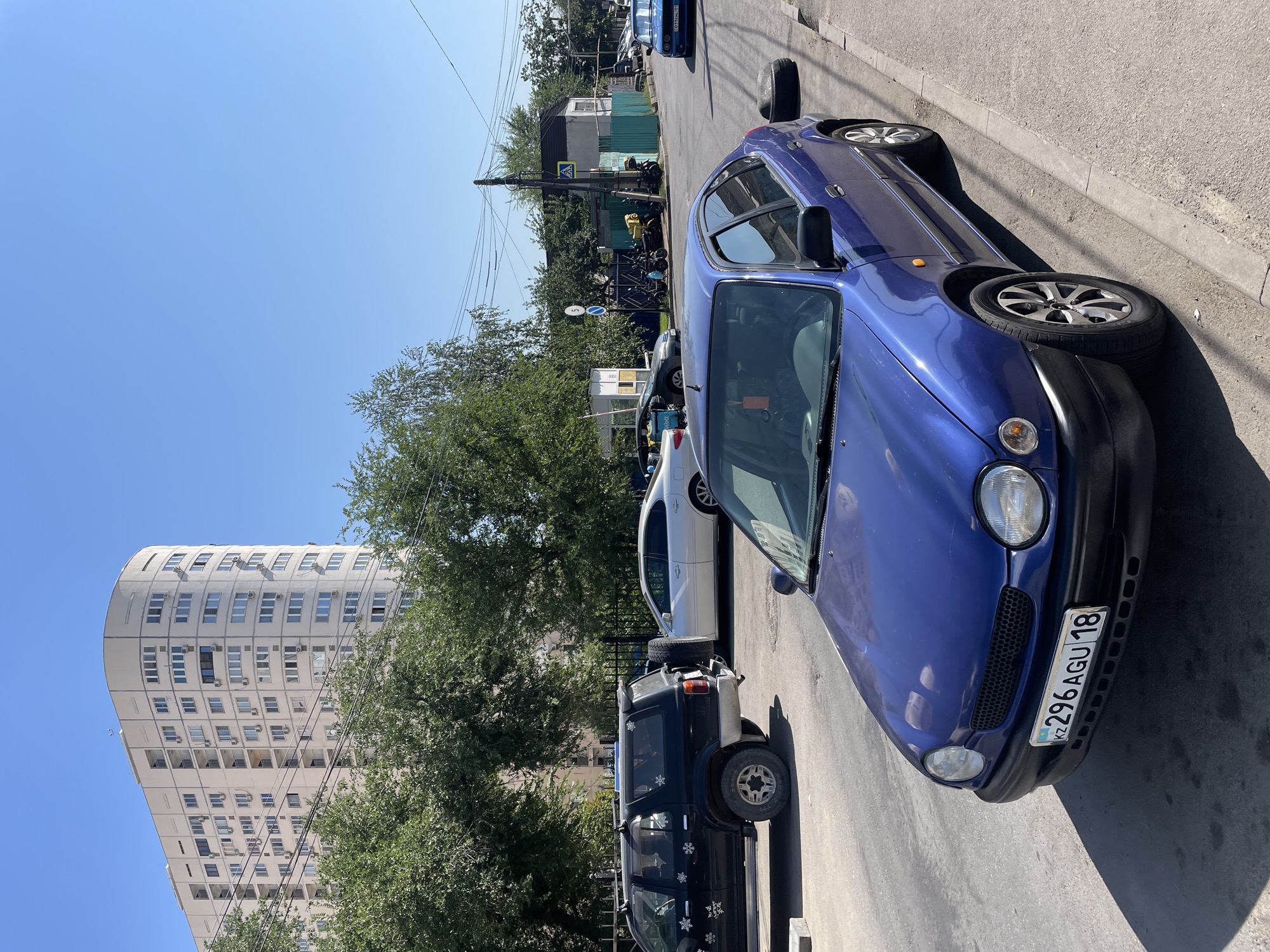 This is a daytime, eye-level, outdoor shot of a residential street in Almaty, Kazakhstan. In the foreground, a blue sedan is parked on the right side of the street, facing away from the camera. The car has a license plate that reads "KZ 296 AGU 18". To the left of the blue car, a black SUV is parked, facing left, with a spare tire mounted on its rear. Further down the street, a white sedan is parked, and behind it, several motorcycles are visible. In the background, a tall, curved, light-colored apartment building stands against a clear blue sky. Trees line the street, and power lines are visible overhead. The scene appears to be a typical urban residential area with parked vehicles and apartment buildings.
