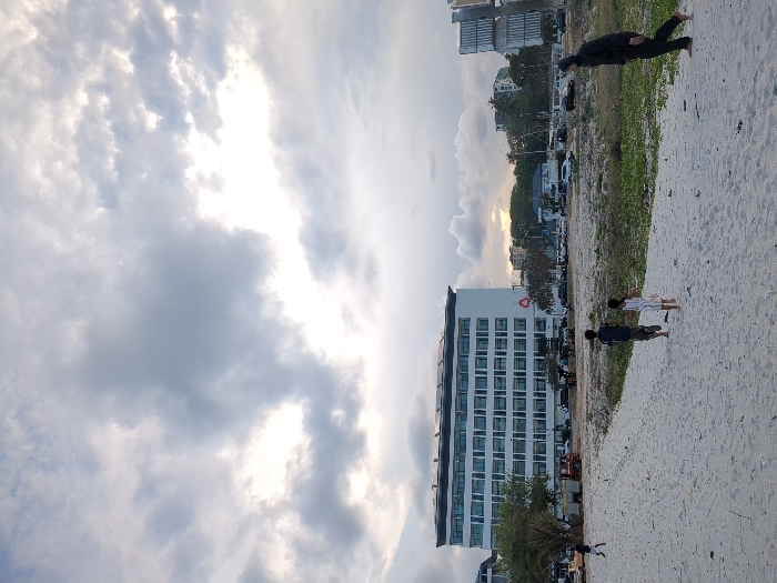 The image shows a white sandy beach with four people on it. The three children are standing on the sand, while an adult male wearing a dark jacket and black pants is walking away from the camera. The background is a cityscape with a large white building in the center, a few other buildings to the right, and a small, dense, green tree to the left. The sky is cloudy with a white sun shining through. This location is on the coast in Dar es Salaam, Tanzania, at coordinates -6.802 latitude and 39.289 longitude.