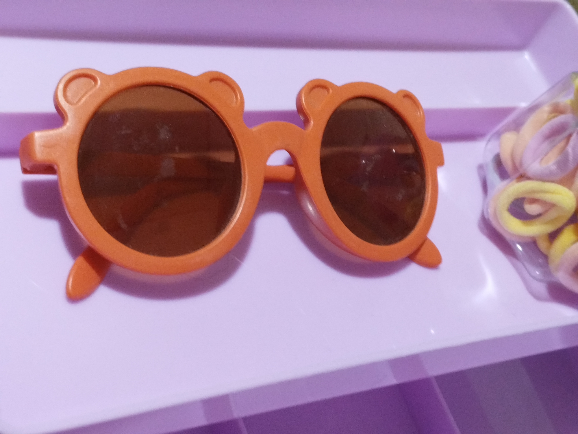 On a light purple surface are a pair of children's sunglasses and a partially visible plastic bag containing hair ties. The sunglasses are orange and styled with small ears on the frames, resembling a bear. The hair ties are pastel colors. The location in the city of Lhokseumawe, Indonesia, is not evident in the image.