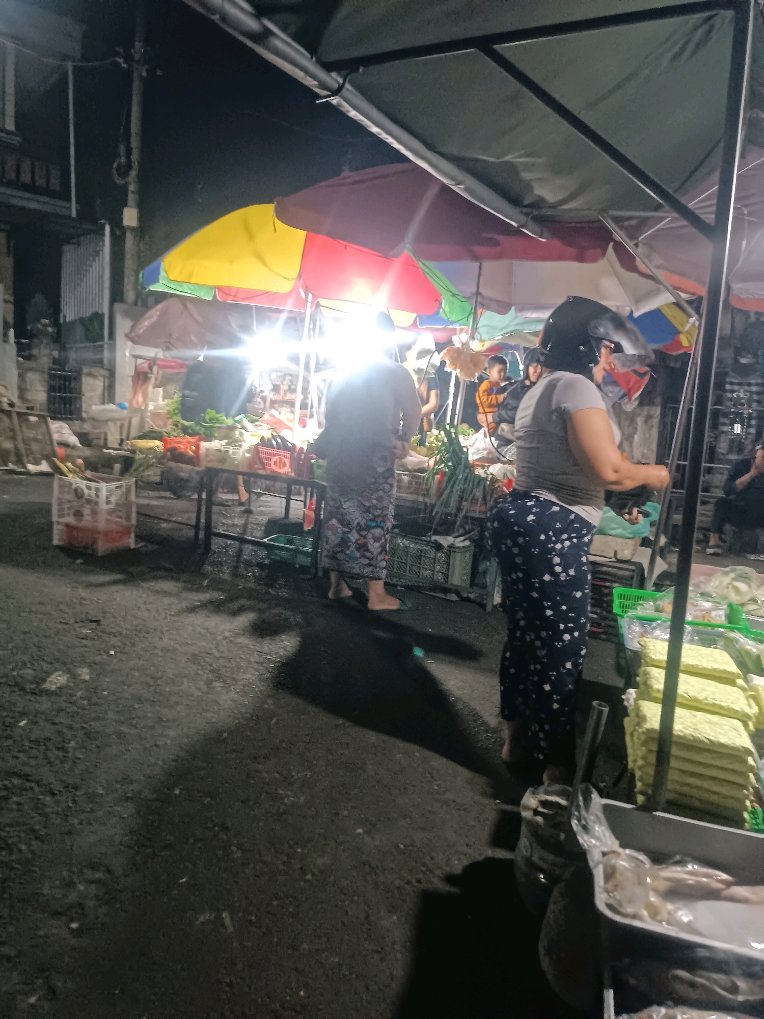 The image depicts a night market scene in Tabanan, Indonesia. The foreground shows a paved surface, likely a street, with dark shadows cast by overhead lighting and structures. A woman, wearing a gray t-shirt and dark, patterned pants, is visible from the waist down, standing near a stall. She has a helmet on, suggesting she may have arrived by motorcycle. To her left, a cart holds a plastic bag filled with fish. In the mid-ground, more stalls are set up under colorful umbrellas, illuminated by bright lights. These stalls appear to be selling various goods, including produce such as tomatoes and leafy greens, displayed in crates. Several other individuals are present in the background, some appearing to be shoppers or vendors. The overall atmosphere is that of a bustling local market at night.