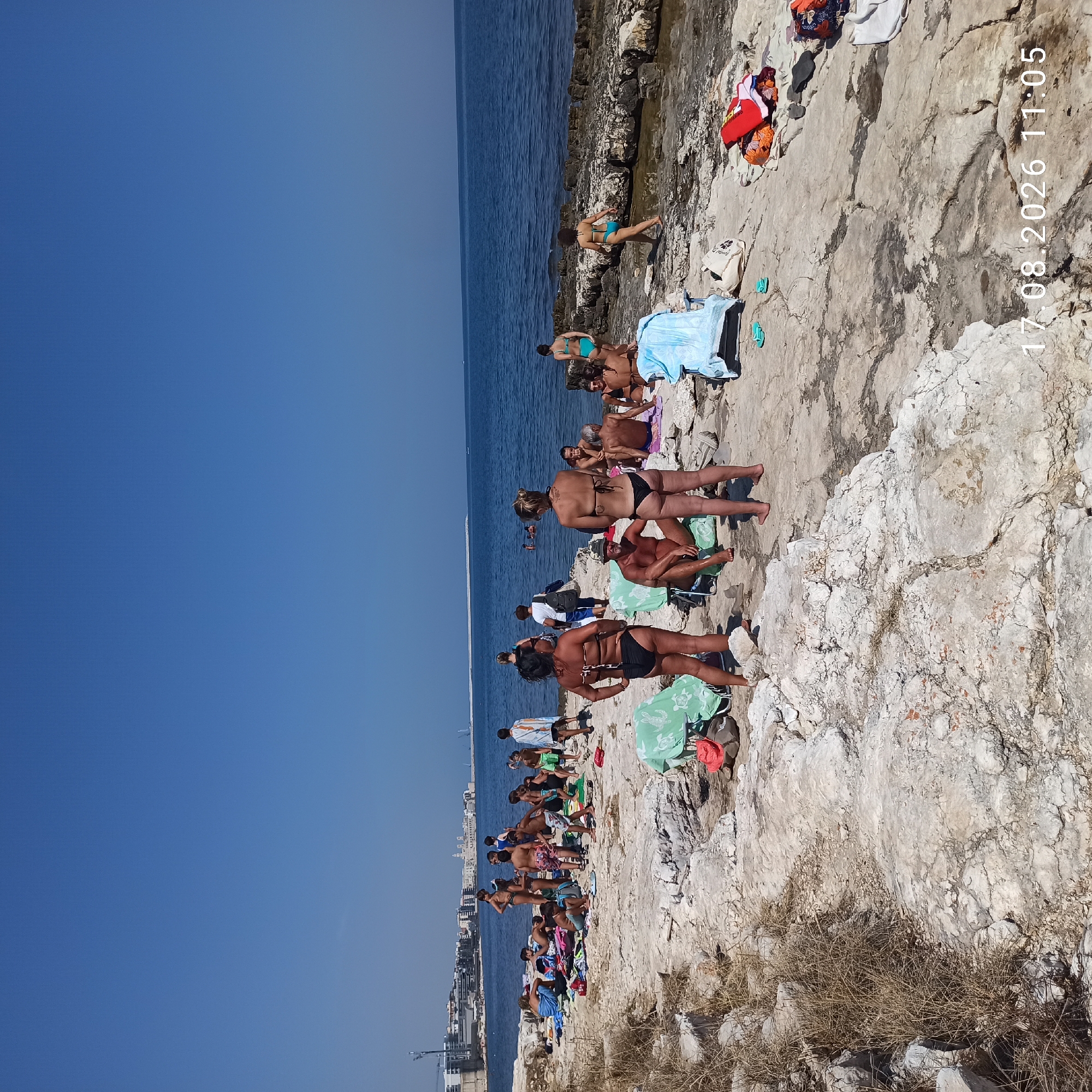 On August 17, 2026, at 11:05 AM, a group of people are sunbathing and relaxing on a rocky coastline in Molfetta, Italy. The scene is bathed in bright sunlight under a clear blue sky. The rocky shore is littered with beach towels, chairs, and bags, indicating a leisurely day at the beach.
In the foreground, several individuals are casually dressed in swimwear, some sitting on towels and others standing. Two women in bikinis are prominently positioned in the center, facing away from the camera. To their left, a man in a hat is seated on a chair, also in swimwear.
Further back, the coastline extends towards the sea, with more people scattered across the rocks. Some are sitting in groups, while others are standing or walking. Children are visible among the adults, suggesting a family-friendly atmosphere.
In the distance, the cityscape of Molfetta can be seen, with buildings and a prominent church steeple dotting the horizon. The blue sea stretches out to meet the sky, with a few distant boats visible. The overall impression is one of a warm, sunny day enjoyed by people at a coastal resort.