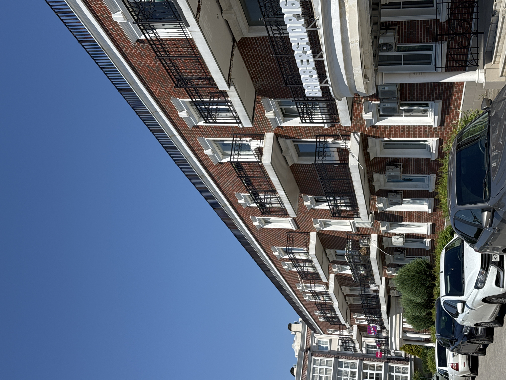 The image shows a row of cars parked in front of a brick building on a sunny day. The building has multiple stories with balconies and windows. A sign that reads "BARBERSHOP IG" is visible on the building. There are trees and bushes in front of the building. The sky is clear blue.