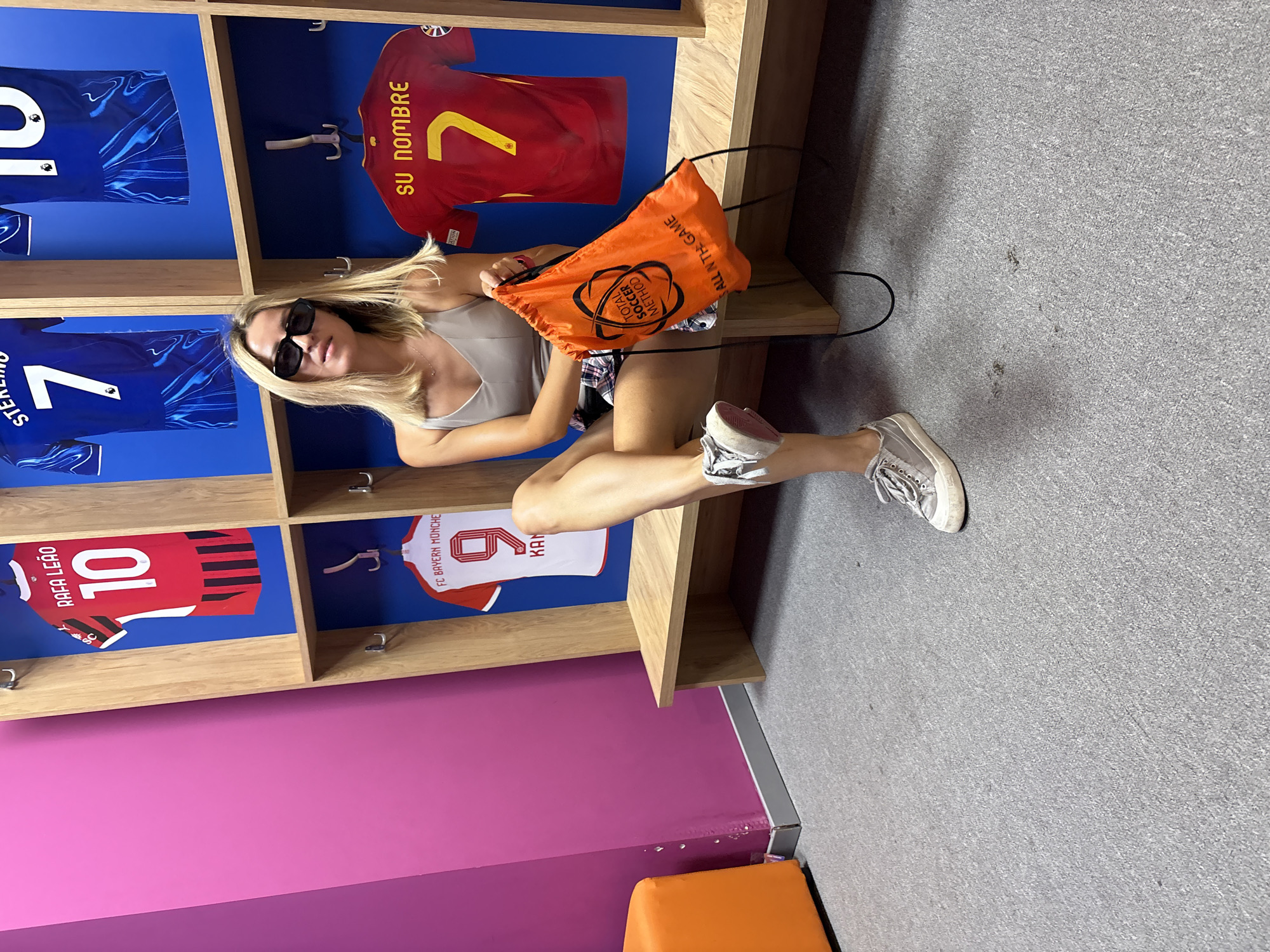 A blonde woman with sunglasses sits on a wooden bench in a locker room with blue walls. She is wearing a grey tank top and plaid shorts, with silver sneakers. In her lap, she holds an orange drawstring bag with "TOTAL SOCCER METHOD" printed on it. Behind her, jerseys hang in cubbies. One red jersey with "RAFA LEÃO 10" and another red jersey with "SU NOMBRE 7" are visible, as well as a white jersey with "FC BAYERN MÜNCHE 9 KAN". The locker room appears to be located in the city of Valencia, Spain, possibly at the Mestalla Stadium. An orange stool is visible in the left of the frame. The floor is carpeted in a grey speckled material.