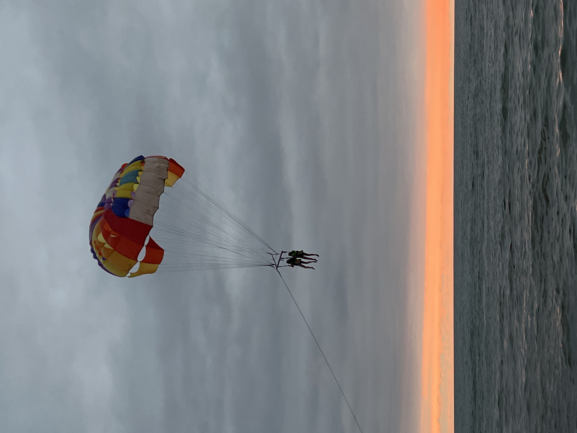 In Ureki, Georgia, at sunset, two people are parasailing over the Black Sea. The parasail is multicolored and vibrant, creating a striking contrast against the overcast sky. The sea is dark and choppy, reflecting the warm hues of the setting sun on the horizon. The scene captures a moment of adventure and relaxation, typical of a beachside resort town.