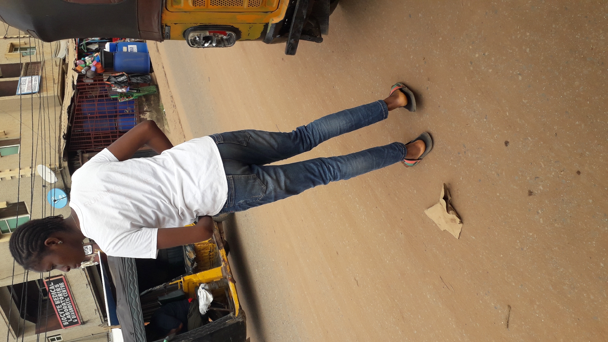 A woman is standing on a street in Nkpor, Nigeria. She is wearing a white t-shirt, blue jeans, and sandals. A yellow vehicle is partially visible in the background. A piece of cardboard lies on the street. Buildings and power lines are visible in the background, along with what appears to be a shop and its contents.
