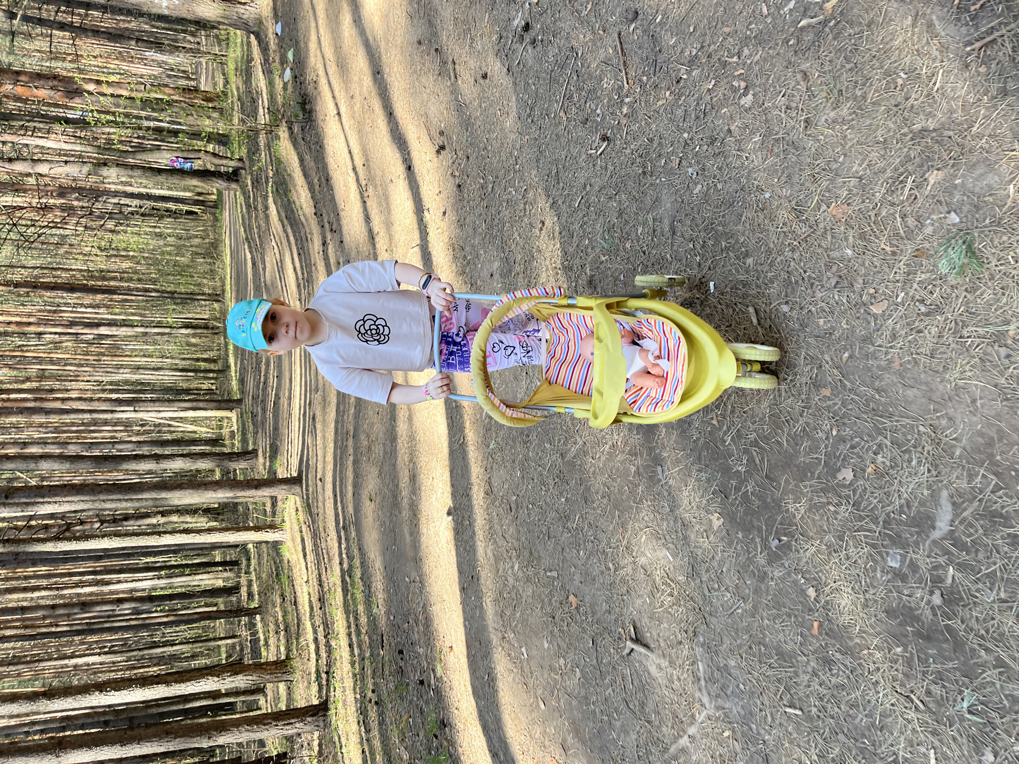 A child wearing a light blue cap and a white long-sleeved shirt with a flower graphic is pushing a yellow stroller containing a doll in a wooded area, most likely in the city of Chaikovsky, Russia. The ground is covered with dirt, dried leaves, and pine needles. Trees are visible in the background. The stroller has a striped lining, and the doll is wearing a similar patterned outfit.