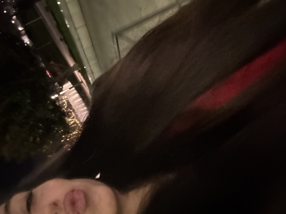 A selfie-style image, dimly lit, features a person's face in the lower left corner, with their tongue sticking out. The background appears to be nighttime outdoors, with decorative lights and possibly a building facade visible. Most of the frame is filled with a dark, blurry mass that could be hair or a garment. A hint of red suggests fabric or lighting. The overall composition is tilted and unbalanced.