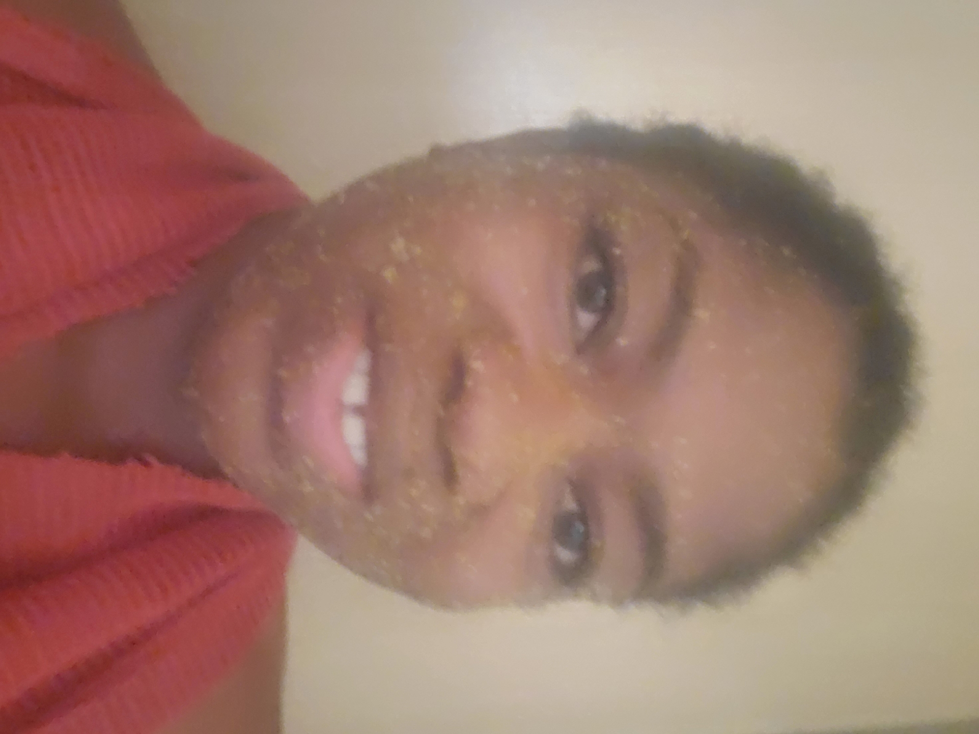 A frontal, close-up portrait depicts an adult individual with dark skin and short, dark, curly hair, smiling directly at the camera. Their face is entirely covered with numerous small, light yellowish-green, granular particles, consistent with a facial scrub or mask. The person's mouth is open in a smile, revealing a visible gap between their two upper front teeth. A red, textured fabric, likely a towel or garment, is draped around their neck and shoulders. The background is a plain, uniformly light beige or off-white wall, indicating an indoor setting. The image provides no visual cues to determine a specific geographic location, such as the city of Kakau, Nigeria.