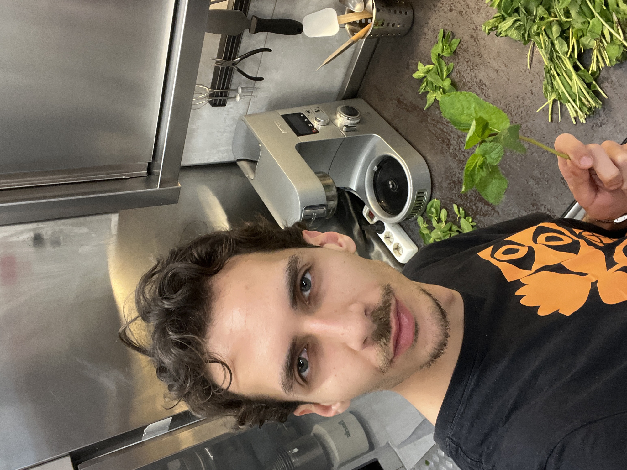A man is in a kitchen, holding a sprig of mint. He has dark curly hair, a mustache, and is wearing a black t-shirt with an orange graphic. Behind him is a stainless-steel food mixer, various kitchen utensils, and a bundle of mint. The location is a kitchen; the city is unknown.