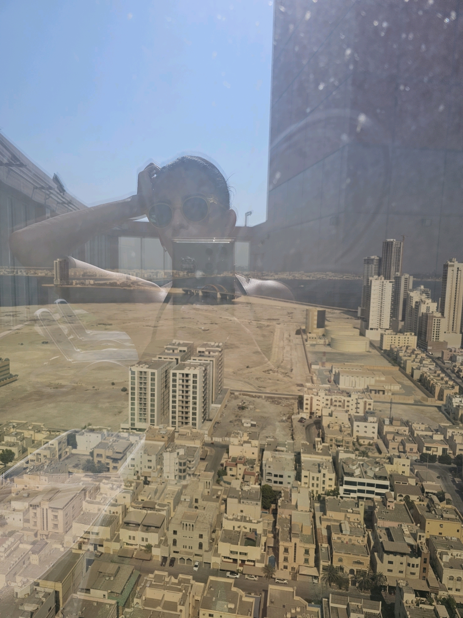 A selfie is taken through a window, reflecting the sky and a person wearing sunglasses and a black top, with their hand on their head. The reflection overlays a view of a city, appearing to be Manama, Bahrain, characterized by a dense urban landscape with a mix of modern high-rise buildings and smaller residential structures. The buildings are predominantly beige and white, with a grid-like street layout visible. In the distance, a body of water can be seen, suggesting a coastal location. The scene is characterized by a bright, sunny day, indicated by the clear blue sky in the reflection. The image is also affected by the window's condition, showing smudges and dust particles that create a slightly hazy and diffused appearance, particularly on the building facades.