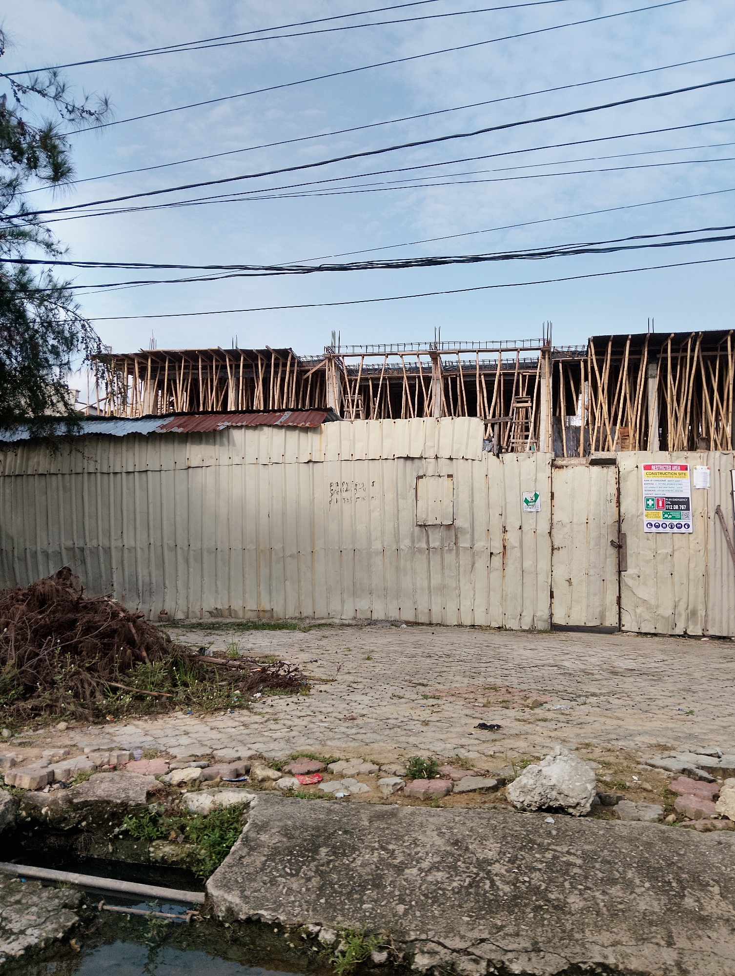 The image depicts a construction site in Moba, Nigeria. A corrugated metal fence, showing signs of rust and wear, encloses the site. Above the fence, the wooden scaffolding of a multi-story building under construction is visible, supported by numerous vertical wooden poles. The sky above is partly cloudy with several power lines strung across it. In the foreground, a pile of debris, consisting of branches and roots, lies on a cobblestone ground. Adjacent to the debris is a narrow channel of murky water, with concrete slabs at its edge. On the metal fence, there are several signs, including one indicating "RESTRICTED AREA CONSTRUCTION SITE" with emergency contact information. There are no people visible in the image.