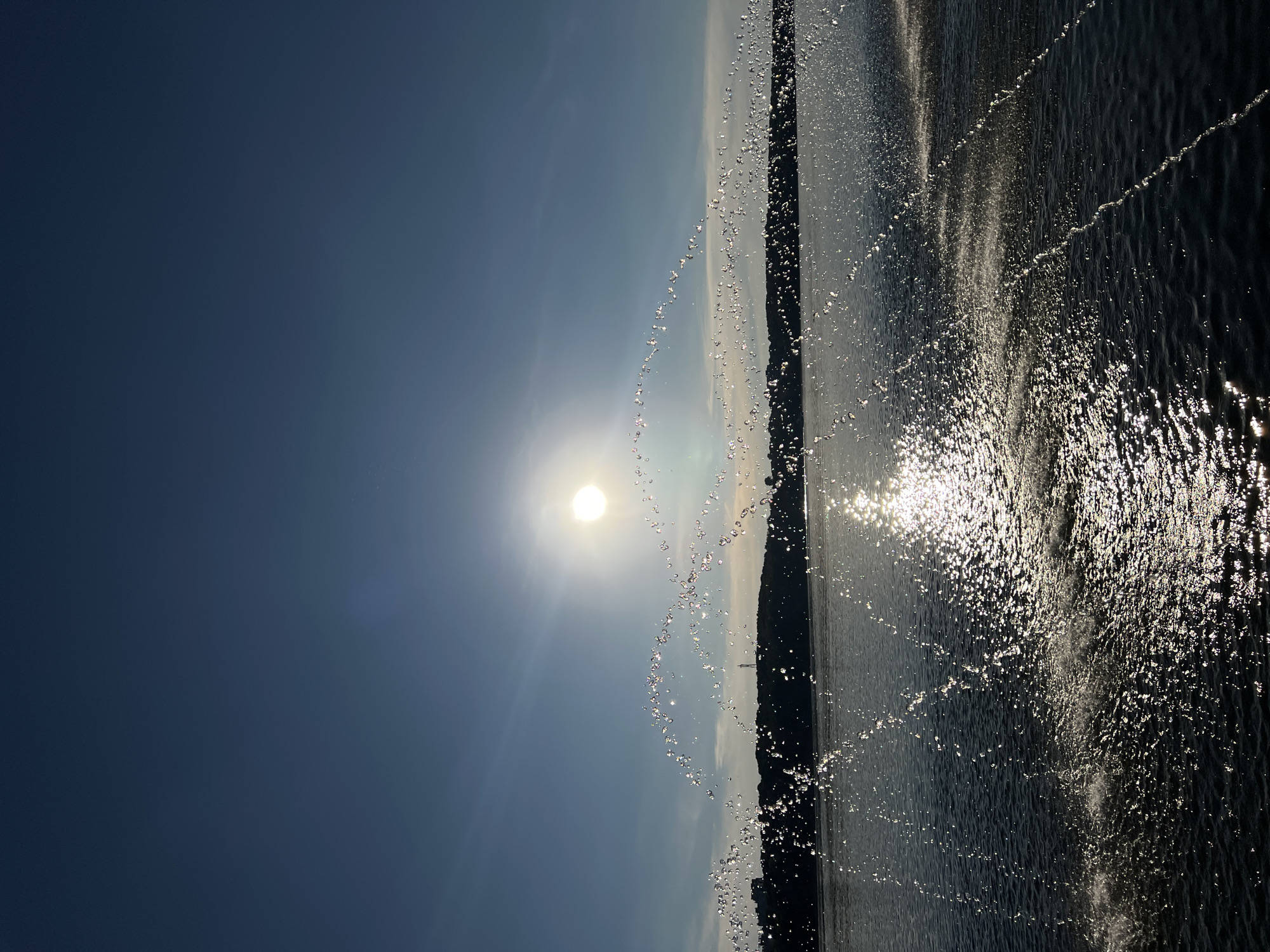 The image depicts a body of water, likely Ternopil Pond, under a clear blue sky with the sun prominently visible. A fountain is in operation, with arcs of water droplets creating a sparkling effect against the sunlight. In the distance, a silhouetted tree line borders the water, with what appears to be a city skyline and a radio tower visible atop the hills. The water itself is dark and reflects the sunlight, with smaller splashes and ripples evident. The overall impression is of a bright, sunny day with a recreational or scenic setting.