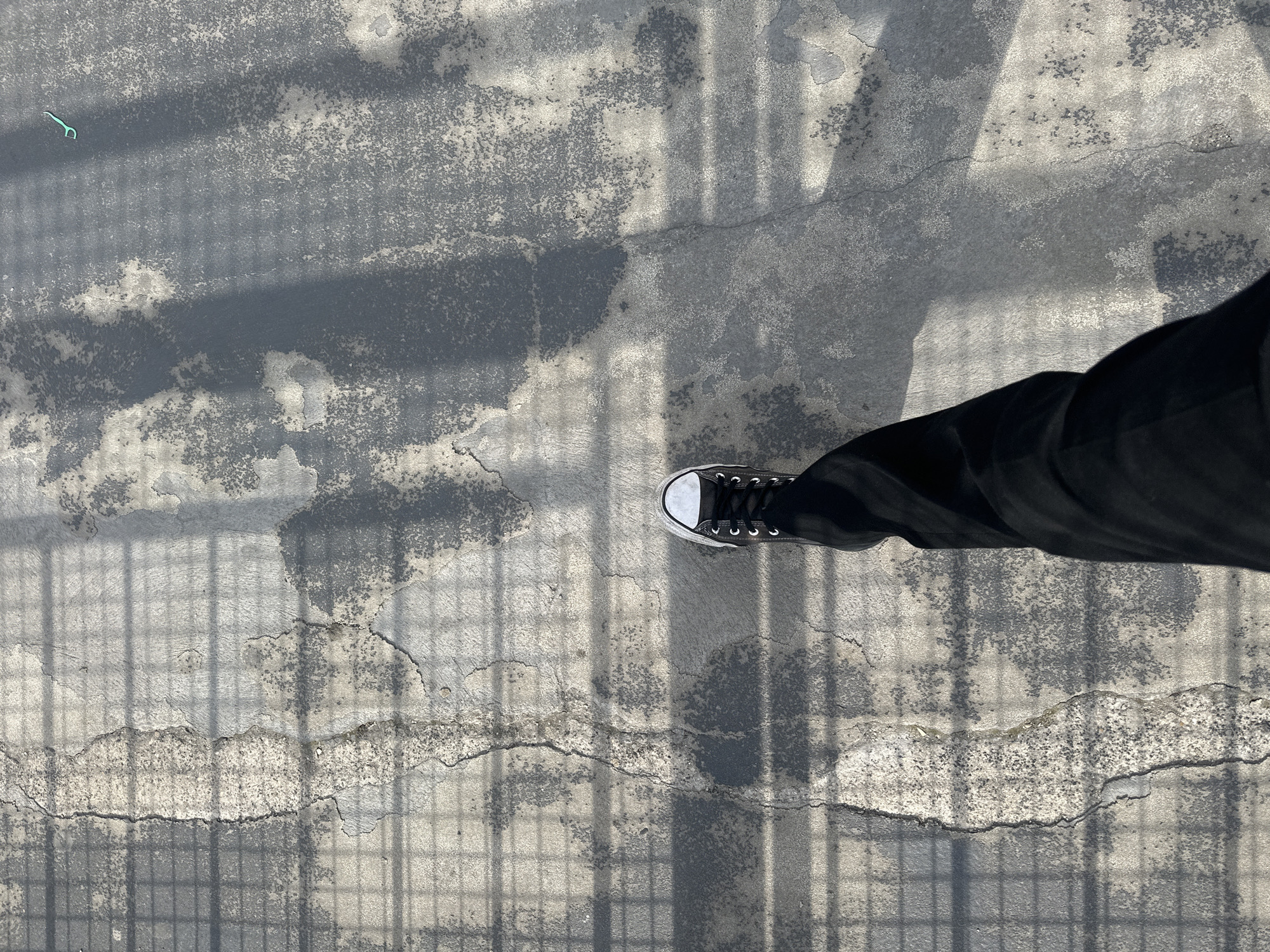 The image depicts a person standing on a cracked, weathered concrete surface in an outdoor location. The person is wearing black pants and a black and white Converse sneaker. The surface is marked by shadows of a wire mesh or grid-like structure, likely a fence or railing above. The background surface appears to be a road or sidewalk. A green dental floss pick is visible in the upper left of the image. No further details are available to indicate that the photo was taken in Nogent-sur-Oise, France.