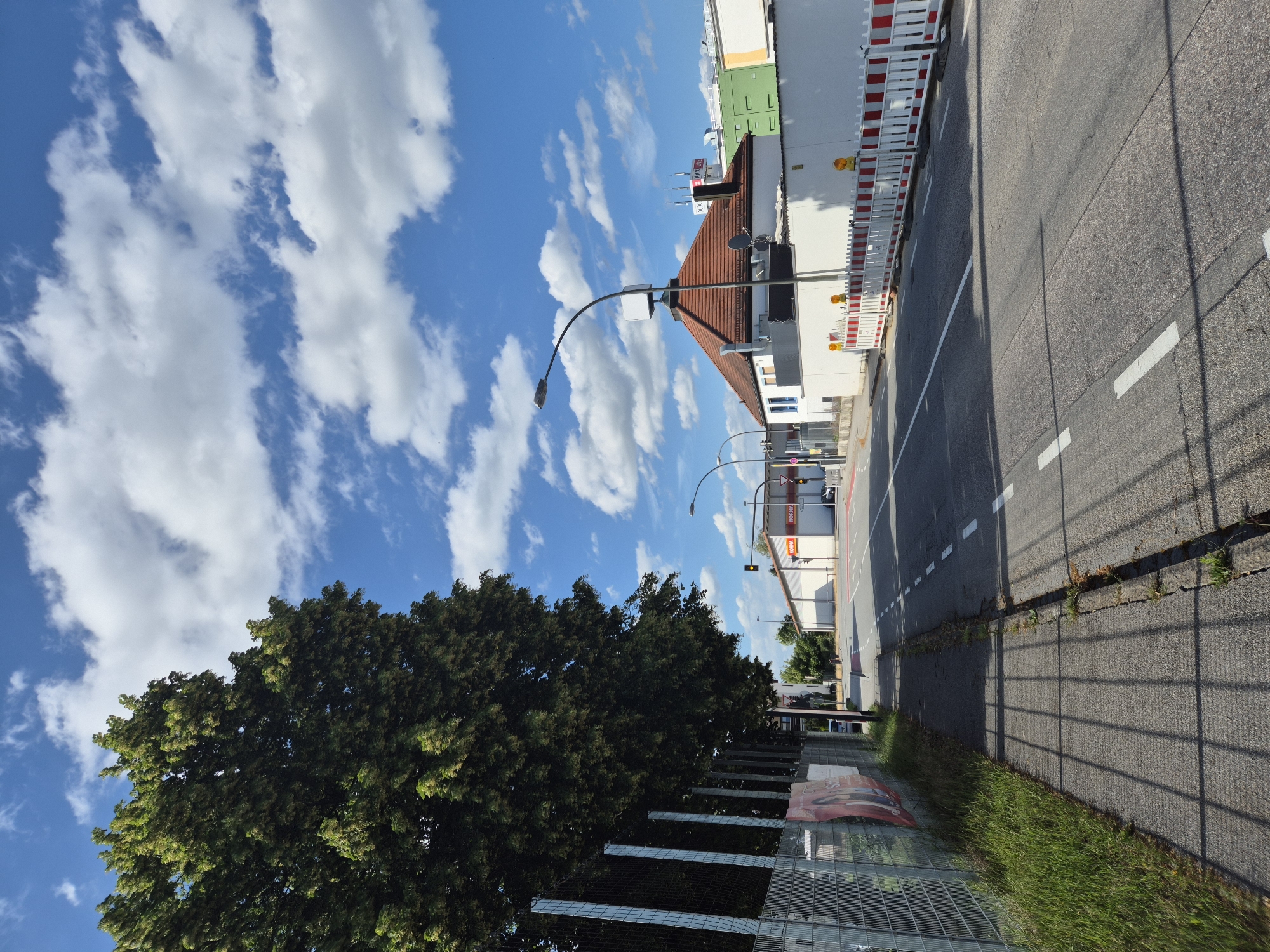 The image depicts a street scene in Landshut, Germany, on a sunny day with scattered clouds. On the left side of the road, a chain-link fence with metal posts runs alongside a grassy area and a sidewalk. A large tree with dense green foliage partially obscures the sky. The road itself is paved and marked with white dashed lines, indicating traffic lanes.
On the right side of the road, several buildings are visible. One building has a prominent red-tiled roof and a white facade, with a street lamp in front of it. Another building, likely a commercial establishment, features the "NORMA" logo on its facade and has a traffic light in front. Further down the street, construction barriers with red and white stripes are set up along the side of the road, suggesting ongoing work. The overall environment appears to be an urban or suburban setting with a mix of residential and commercial properties. There are no people visible in the image.