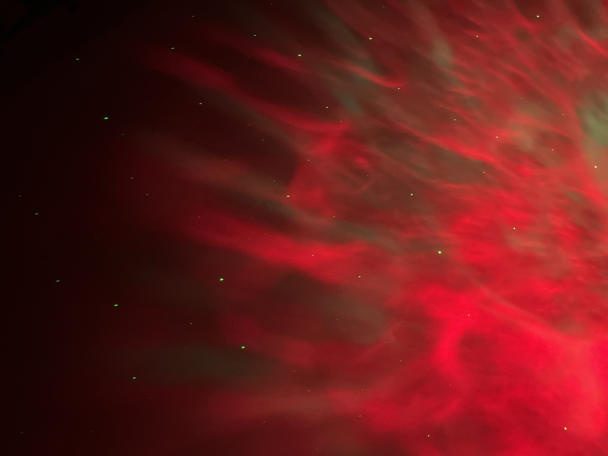 The image depicts a dark, atmospheric scene with swirling patterns of red and green light, reminiscent of nebulae or aurora borealis. Numerous small, bright green dots are scattered across the frame, appearing like distant stars or light particles. The lighting suggests a theatrical or artificial environment, possibly created by projectors or lasers. There are no people, identifiable objects, or environmental features that would place this image in a specific location within the city of Saint Petersburg, Russia. The abstract nature of the image makes it impossible to ascertain a specific context or setting within the city.