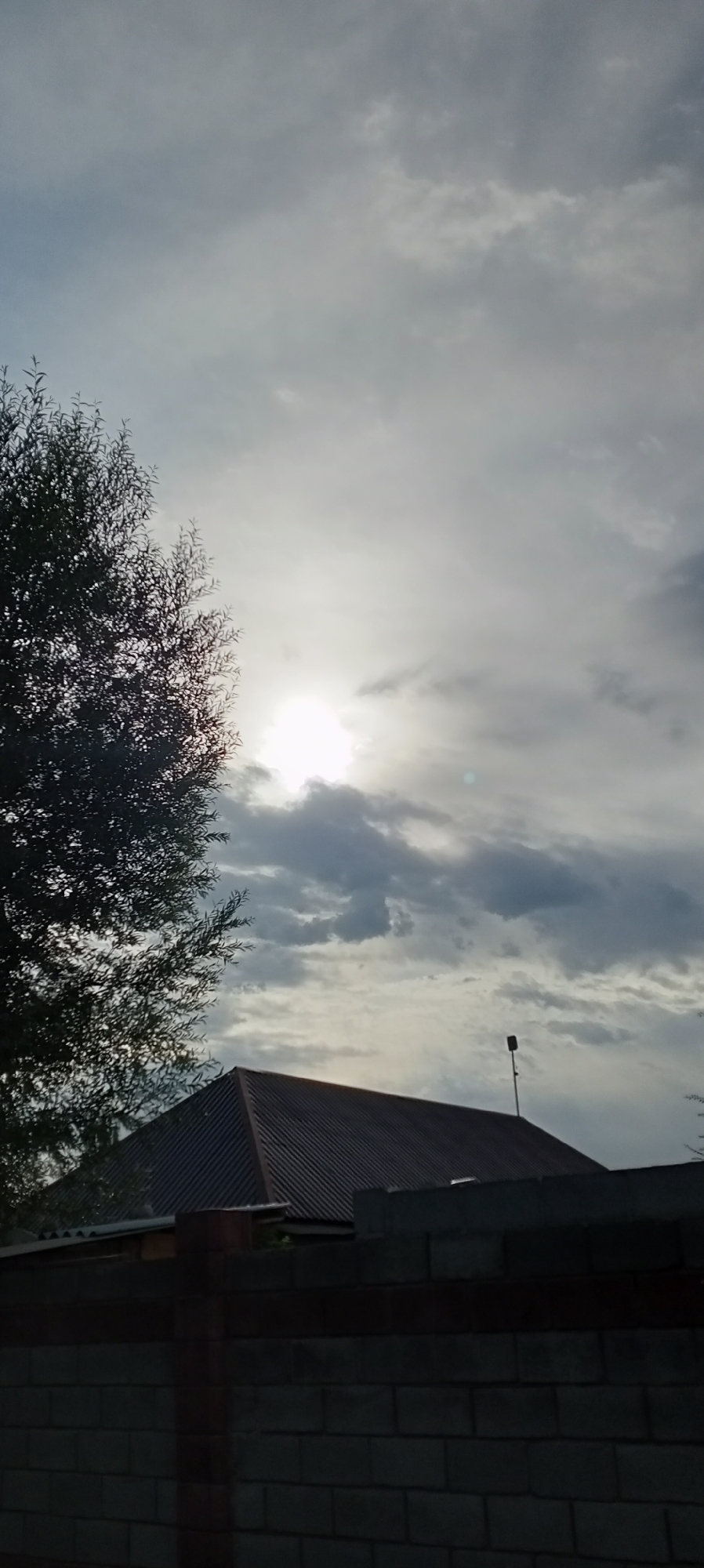 The image depicts an overcast sky with the sun partially visible through the clouds, casting a soft glow. In the foreground, a silhouette of a tree with sparse leaves is seen on the left. Below the sky, the dark, sloped roof of a building is visible, with a dark brick fence in the lower portion of the frame. The scene appears to be taken in an outdoor environment, possibly a residential area in Sokuluk, Kyrgyzstan, given the architectural style of the roof and fence. No people or specific actions are discernible in the image.