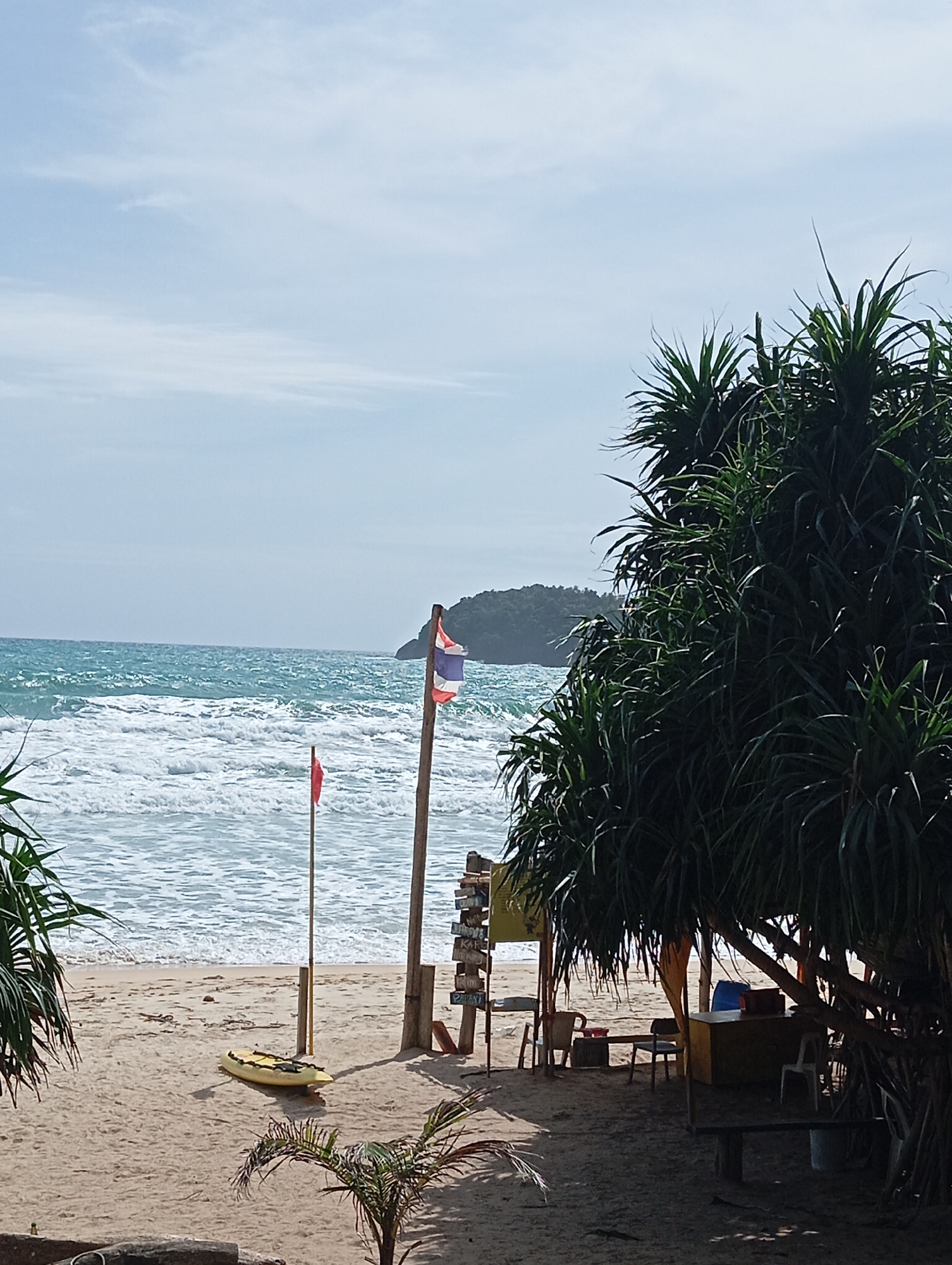 The image depicts a serene beach scene in Karon, Thailand. A sandy beach stretches towards the turquoise ocean, where gentle waves create a foamy white crest. The sky above is a soft blue with scattered wisps of clouds.
On the beach, a yellow kayak rests near the water's edge, a red flag planted in the sand beside it. In the background, a Thai flag flutters on a wooden pole, standing sentinel against the backdrop of a distant, lush green island.
To the right, a cluster of large, spiky palm-like trees casts a dappled shade over a small seating area. A rustic wooden table is set with chairs, suggesting a place for visitors to relax and enjoy the coastal view. A low wooden bench is also visible. Several signs, possibly indicating directions or services, are mounted on a wooden post near the trees. The overall atmosphere is tranquil and inviting, capturing the essence of a peaceful day at a tropical beach.