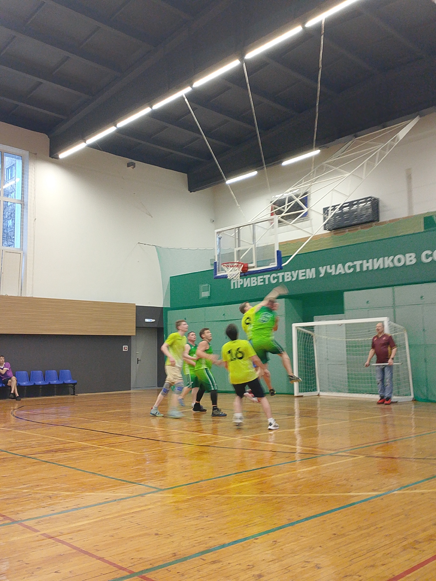 The image shows a basketball game in progress in what appears to be a gymnasium. Several players in green and yellow uniforms are actively engaged in play near a basketball hoop. One player is airborne, presumably attempting a shot or dunk. Another player is wearing number 16 on his jersey. A man in a maroon shirt and jeans stands on the sidelines, possibly a coach or referee, holding a white object. In the background, there is a set of blue chairs occupied by one person. A sign above the basketball hoop features Cyrillic script, which translates to "Welcome participants." Based on the sign's language and location data, the event is likely occurring in Izhevsk, Russia. The gymnasium has a high ceiling with industrial lighting.