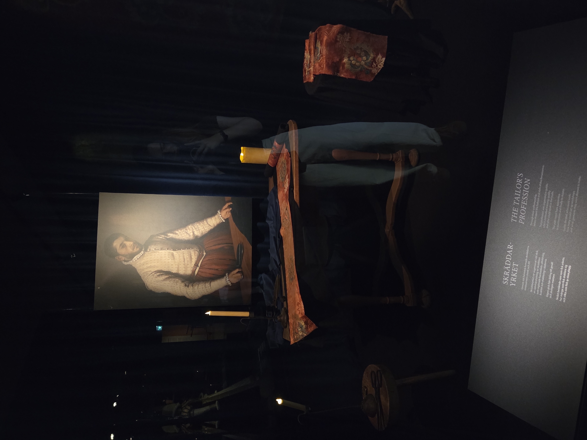 The image depicts an exhibit focused on tailoring, likely displayed within a museum. In the background, a large portrait shows a man dressed in Renaissance-era clothing, featuring a patterned tunic and a red skirt. In the foreground, a wooden table is set up with tailoring tools, including scissors, and draped with fabric. Two lit candles provide illumination. A person, seen in reflection, is holding a phone and taking a picture of the exhibit.
The exhibit includes a descriptive plaque at the bottom of the image, which reads "SKRÄDDAR-YRKE T" in Swedish and "THE TAILOR'S PROFESSION" in English. This indicates the exhibit is located in Malmö, Sweden, as evidenced by the Swedish text. The dark background and the staged presentation suggest it is an indoor exhibition, possibly at a museum like Malmö Museer.
