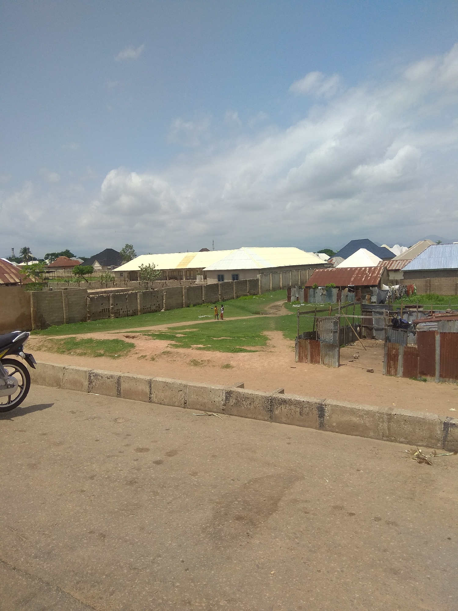 This media file depicts a sunny day in Jalingo, Nigeria, with a view of a residential area. In the foreground, a motorcycle is partially visible on the left side, parked on a paved road. The road is bordered by a concrete curb.
Beyond the curb, there is a patch of dirt and then a grassy area. A long, light-colored building with a large roof, possibly a school or community center, dominates the middle ground. It is enclosed by a concrete wall. To the right of this building, there are several smaller structures made of corrugated metal, appearing to be makeshift homes or stalls.
Two young boys are visible in the grassy area, dressed in what appear to be school uniforms, suggesting they might be playing or socializing. In the background, various houses with different roof types, including corrugated metal and tiled roofs, are scattered across the landscape. Some trees are interspersed among the buildings. The sky is partly cloudy, with white clouds against a clear blue sky, indicating it is likely daytime, possibly late morning or early afternoon. The overall impression is of a typical urban or semi-urban environment in Nigeria. No visible text is present in the image.