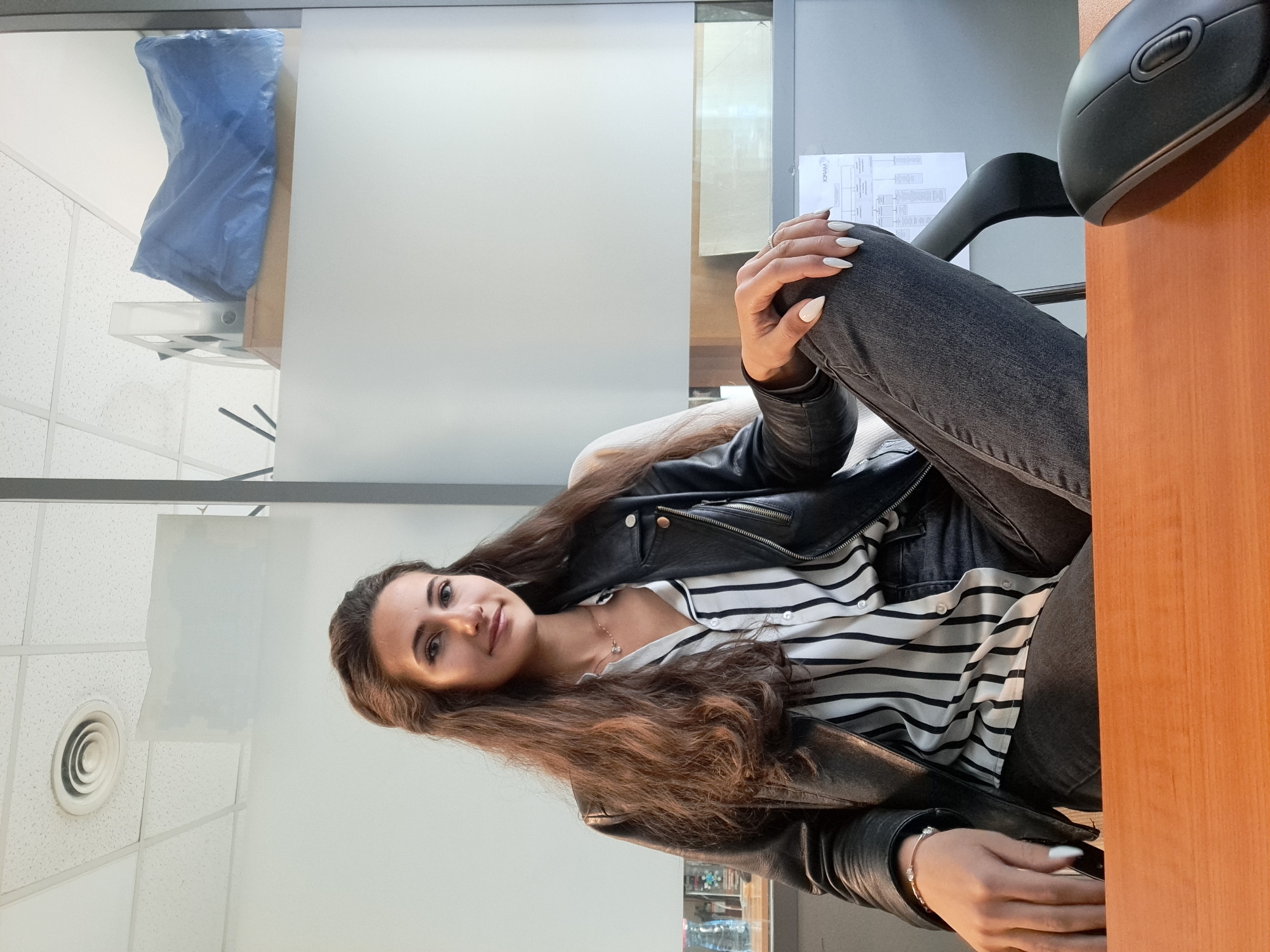 A young woman with long, brown hair is sitting at a wooden desk in an office in Sofia, Bulgaria. She is wearing a striped shirt, black jeans, and a black leather jacket. She has long white fingernails. She is sitting with her legs crossed and her hands resting on her knee. A computer mouse is visible on the desk near her right arm. A document is posted on the wall behind her. A blue object is covered with plastic on top of a cubicle. The ceiling is white with a round air vent.