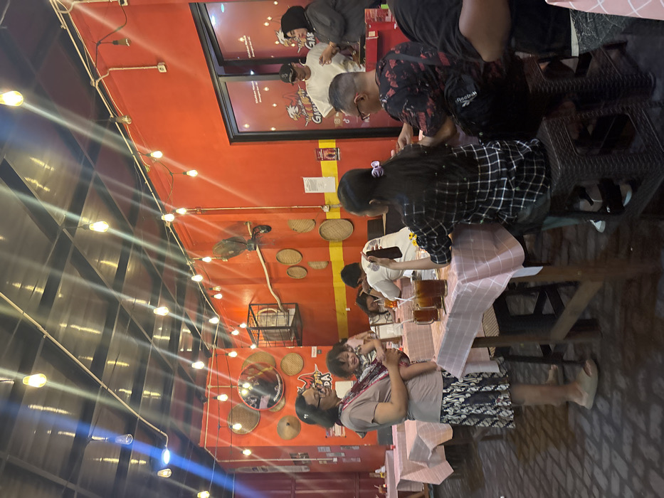 The image captures an evening scene inside a casual restaurant, identified as "Garasi Sidoarjo - Restoran Ayam Bakar," located in Sidoarjo, Indonesia. The establishment features vibrant orange-red walls, decorated with multiple circular woven bamboo winnowing trays of varying sizes and a large mirror. A dark ceiling is illuminated by numerous exposed Edison-style light bulbs strung in rows, casting visible light streaks. The floor is composed of light-colored brick pavers.
In the left foreground, a female subject wearing glasses, a light brown short-sleeved top, and a patterned knee-length skirt stands holding a small child (toddler) in a patterned baby carrier. Both wear light-colored footwear. In the mid-ground, a group of individuals is seated at a square wooden table draped with a pink and white checkered tablecloth. Three glasses containing a dark liquid and straws are visible on this table. One person, wearing a black and white plaid shirt, is seated with their back to the camera. Another male, wearing a black and red patterned shirt and glasses, is seated facing left, with a black Reebok bag visible on his lap. Further back at an adjacent table, a person in a white long-sleeved shirt is partially visible. In the background, behind a glass partition featuring a red chicken logo and "Garasi" text, two individuals are standing: a male wearing a white t-shirt, black baseball cap, and sunglasses, and a female wearing a dark hijab. A wall-mounted fan is situated on the right wall. Exposed electrical conduits and wiring are visible throughout the space.