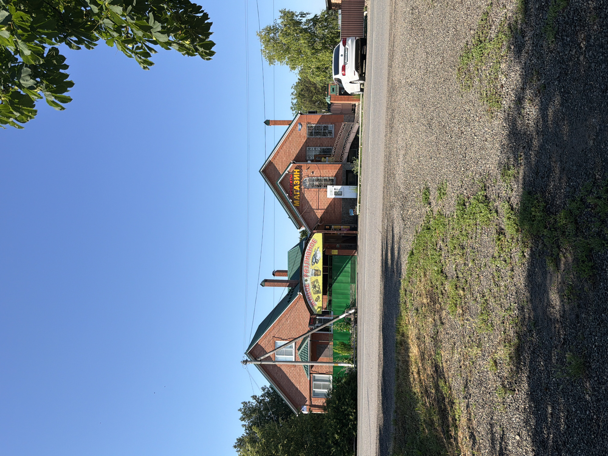 The image shows a sunny day scene in front of two adjacent brick buildings on a street in Leningradskaya, Russia. The building on the left has a green metal gate and a sign that reads "ЖИВОЕ • РАЗЛИВНОЕ" with images of fish. The building on the right has a sign that says "ПРОДУКТЫ МАГАЗИН". A white car is parked in front of the building on the right, and a white vertical machine is next to the entrance of the building on the right. Trees and a fence are visible in the background. The sky is clear blue with a few distant birds. The ground in the foreground is covered with gravel and sparse grass.