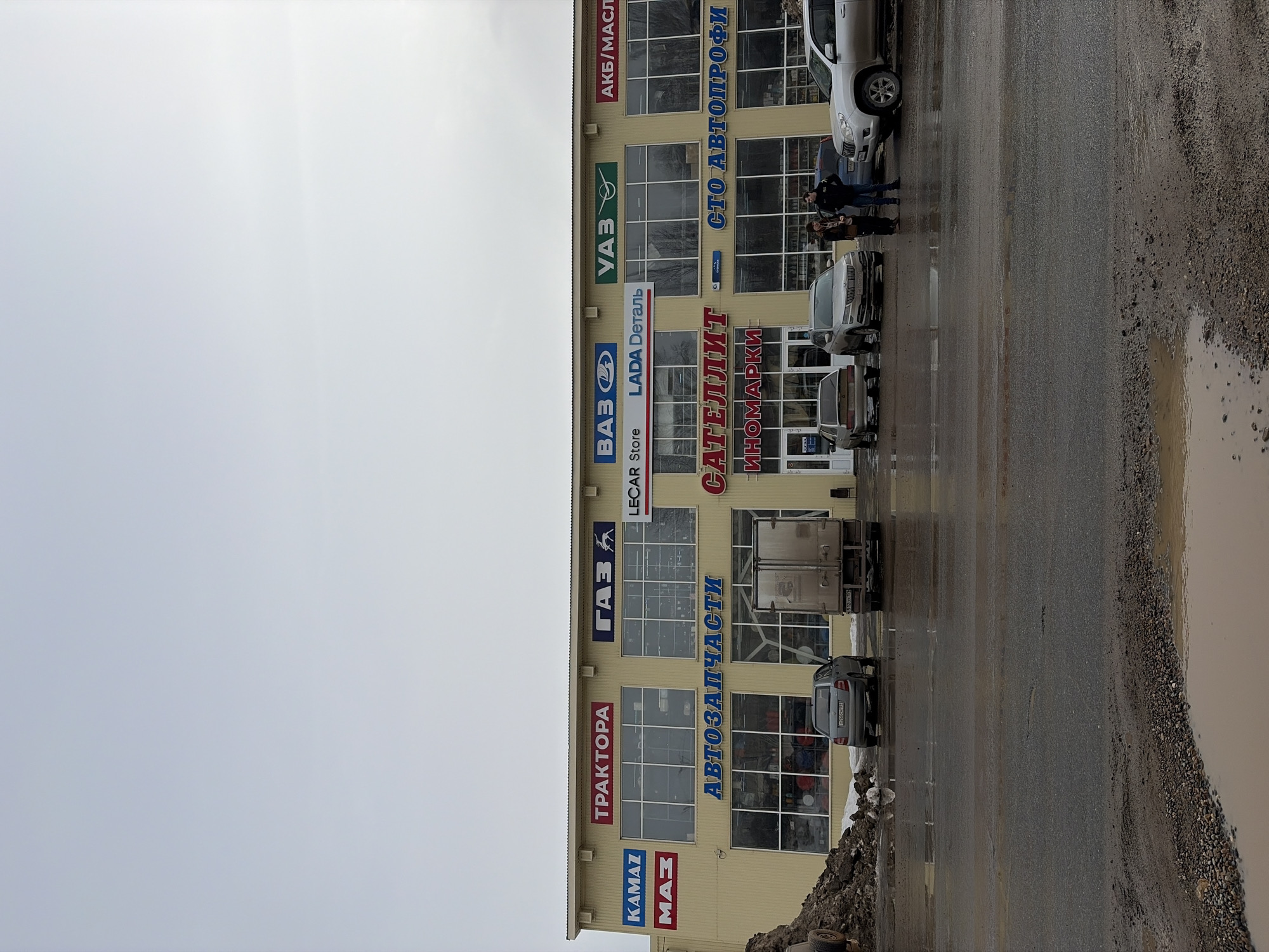 The image depicts a commercial building in Chaikovsky, Russia, under an overcast sky. The two-story structure features a light yellow/beige facade with multiple large, rectangular windows on both levels. Numerous signs in Cyrillic script and Latin characters are affixed to the building, identifying various automotive services and brands. These include "ТРАКТОРА" (Tractors), "ГАЗ" (GAZ), "ВАЗ" (VAZ), "УАЗ" (UAZ), "КАМАZ" (KAMAZ), "МАЗ" (MAZ), "АВТОЗАПЧАСТИ" (Auto Parts), "САТЕЛЛИТ ИНОМАРКИ" (Satellite Foreign Cars), "СТО АВТОПРОФИ" (Service Station Autoprofi), "AKБ/МАСЛА" (Batteries/Oils), "LECAR Store," and "LADA Деталь."
In the foreground, a dark, wet paved area with visible puddles and tire marks extends from the bottom of the frame to the building. A section of gravel and dirt is visible in the immediate foreground, also with small puddles. To the far left, a large pile of dark brown earth mixed with patches of snow or ice is present.
Several vehicles are parked in front of the building. From left to right: a silver Skoda Fabia hatchback (Russian license plate O266OM 193), a light-colored box truck (Russian license plate T532OY 193), a light brown/gold sedan, a silver Skoda Kodiaq SUV (Russian license plate H649BX 193), a partially obscured dark sedan, and a silver Toyota Land Cruiser or similar SUV.
Two individuals are standing on the paved area, positioned between the silver Skoda Kodiaq and the dark sedan. One individual wears a dark jacket and light pants, facing towards the right. The second individual, wearing a dark jacket, faces the first person. Their interaction involves standing in close proximity. The overall scene suggests a commercial area dedicated to vehicle sales and service, likely during a cool or cold season given the puddles and earth pile.
