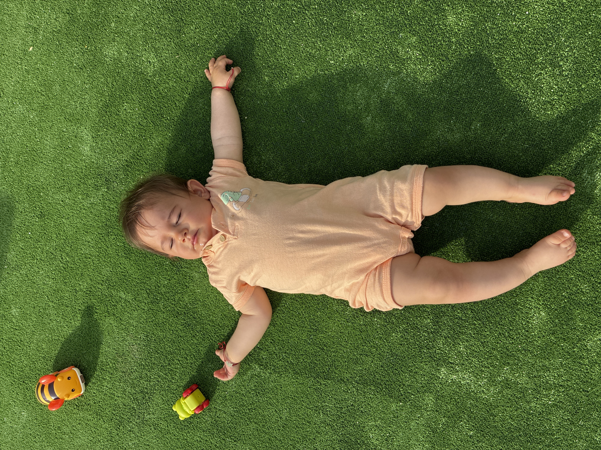 A top-down shot captures a baby sleeping peacefully on bright green artificial turf under clear daylight in Varna, Bulgaria. The infant is wearing a light peach-colored short-sleeved and short-legged romper, which features a collar with three buttons and a small light green dinosaur graphic on the left chest area. A thin red string bracelet is visible on the baby's right wrist. The baby lies on their back with arms outstretched, hands slightly curled, and eyes closed, suggesting a calm and serene moment of rest.
Two colorful plastic toy cars are present on the turf near the baby's left side: one yellow and black toy resembling a bee, and a smaller green and yellow toy. Strong, distinct shadows are cast on the turf by the baby's head, right arm, and the toys, indicating bright sunlight, likely during the morning or afternoon. The texture of the artificial grass is clearly visible. The overall impression is one of tranquility and comfort. No visible text is present in the image.