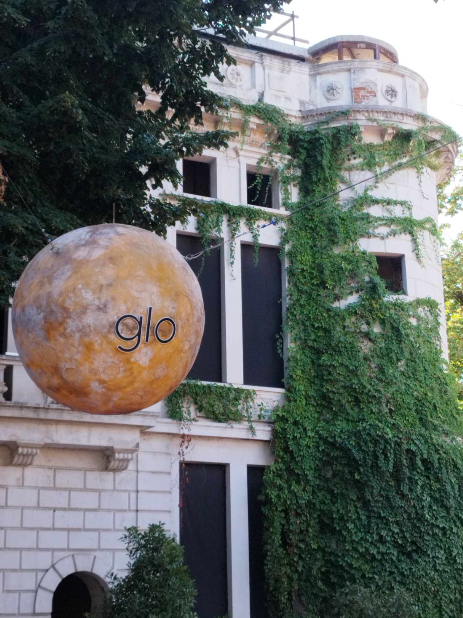 The image depicts the exterior of a building in Bucharest, Romania, featuring a large, spherical sign that reads "glo". The building is partially covered in lush green ivy, and there are several dark-colored windows visible. The sign, which appears to be made of a textured, orange-brown material resembling a celestial body, is suspended from the building by wires. The architecture suggests an older, possibly ornate style, with decorative elements on the facade. The surrounding environment includes trees and foliage, indicating an outdoor setting, likely in a part of the city with historical architecture and greenery.